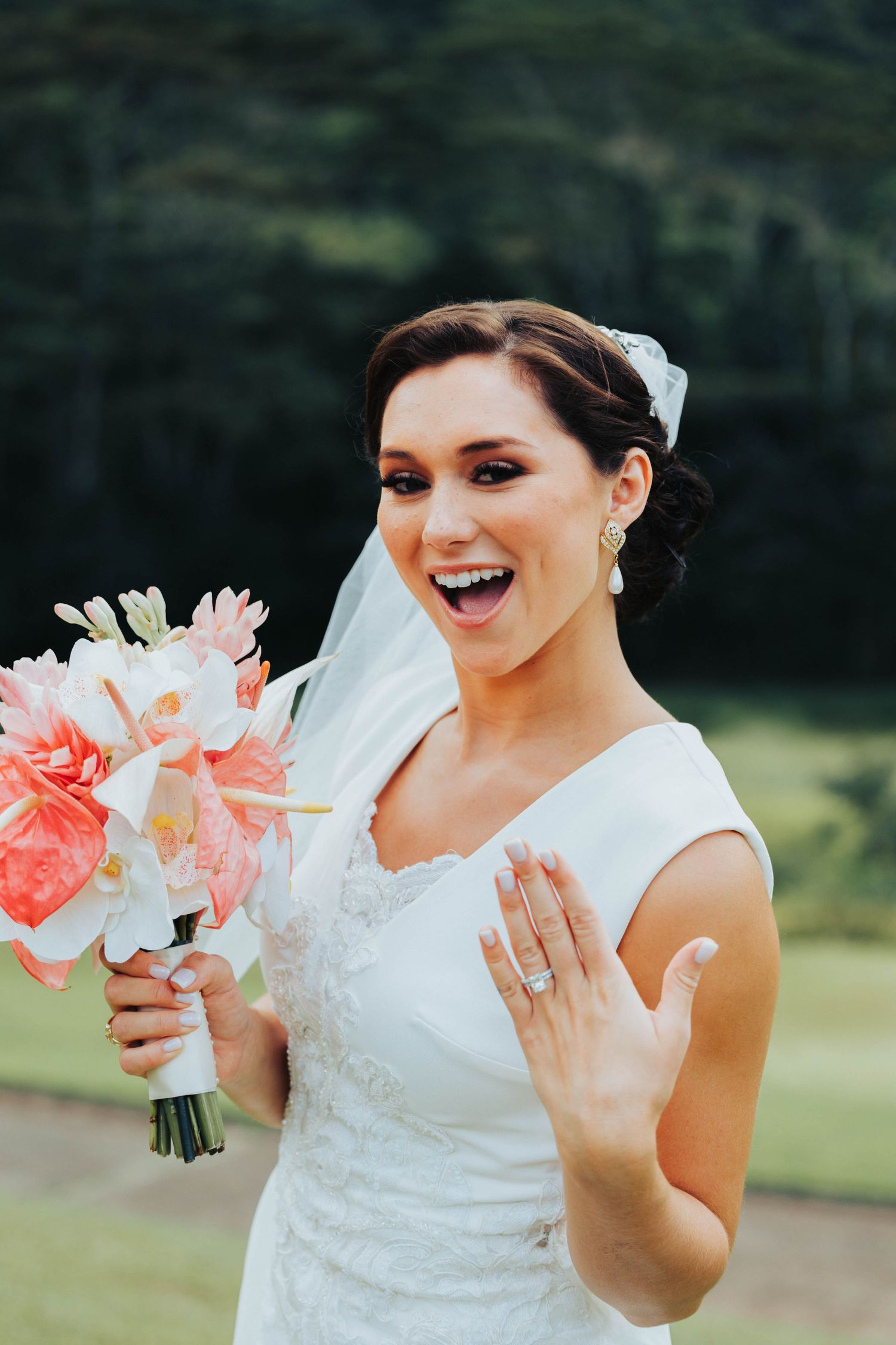 A joyful bride in a white wedding dress laughs while holding a pink and white floral bouquet outdoors.
