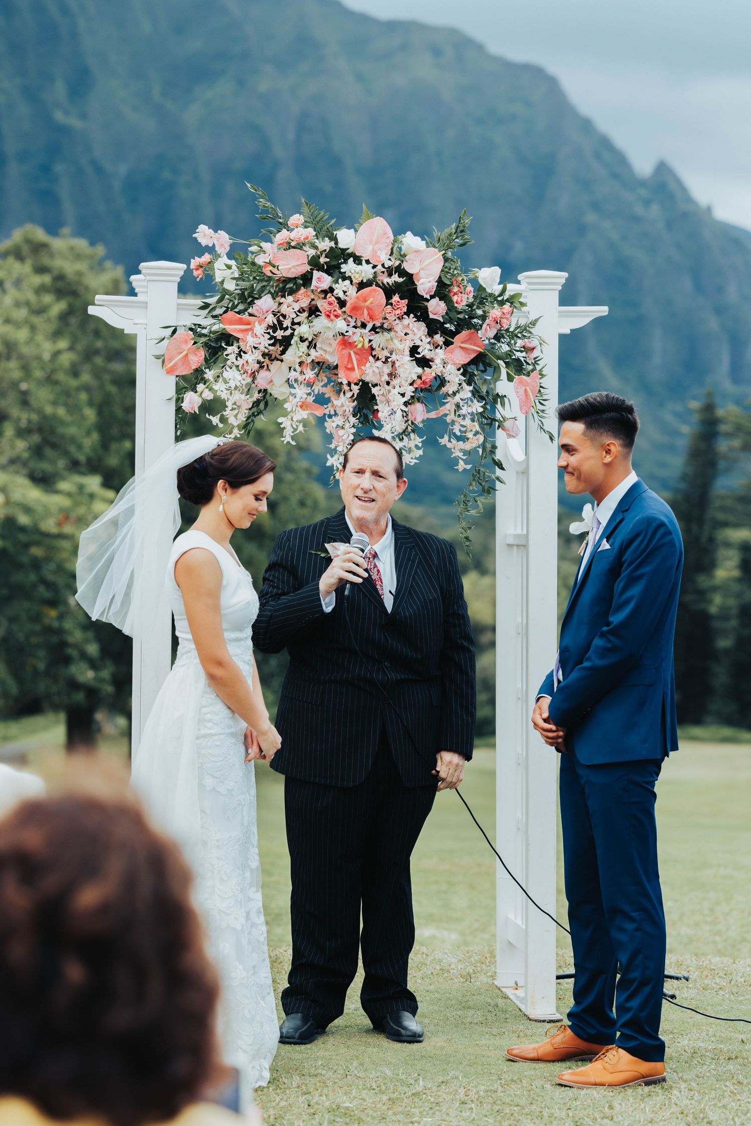 Wedding ceremony taking place outdoors with mountain backdrop and white floral arch decorations.
