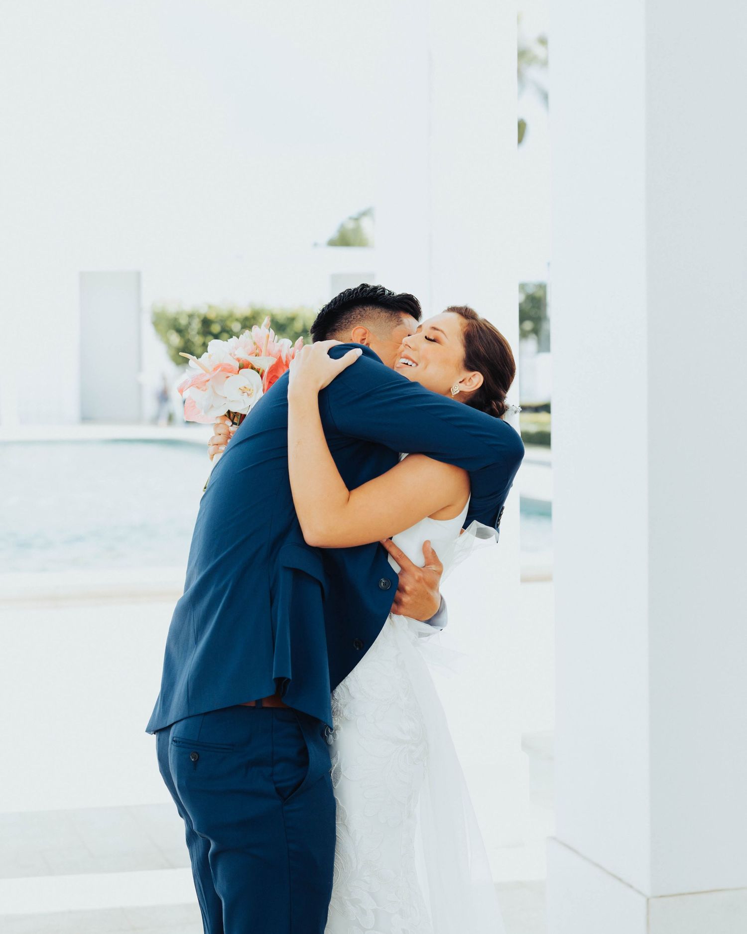 A couple in formal attire share an intimate embrace outdoors against a bright white architectural backdrop.