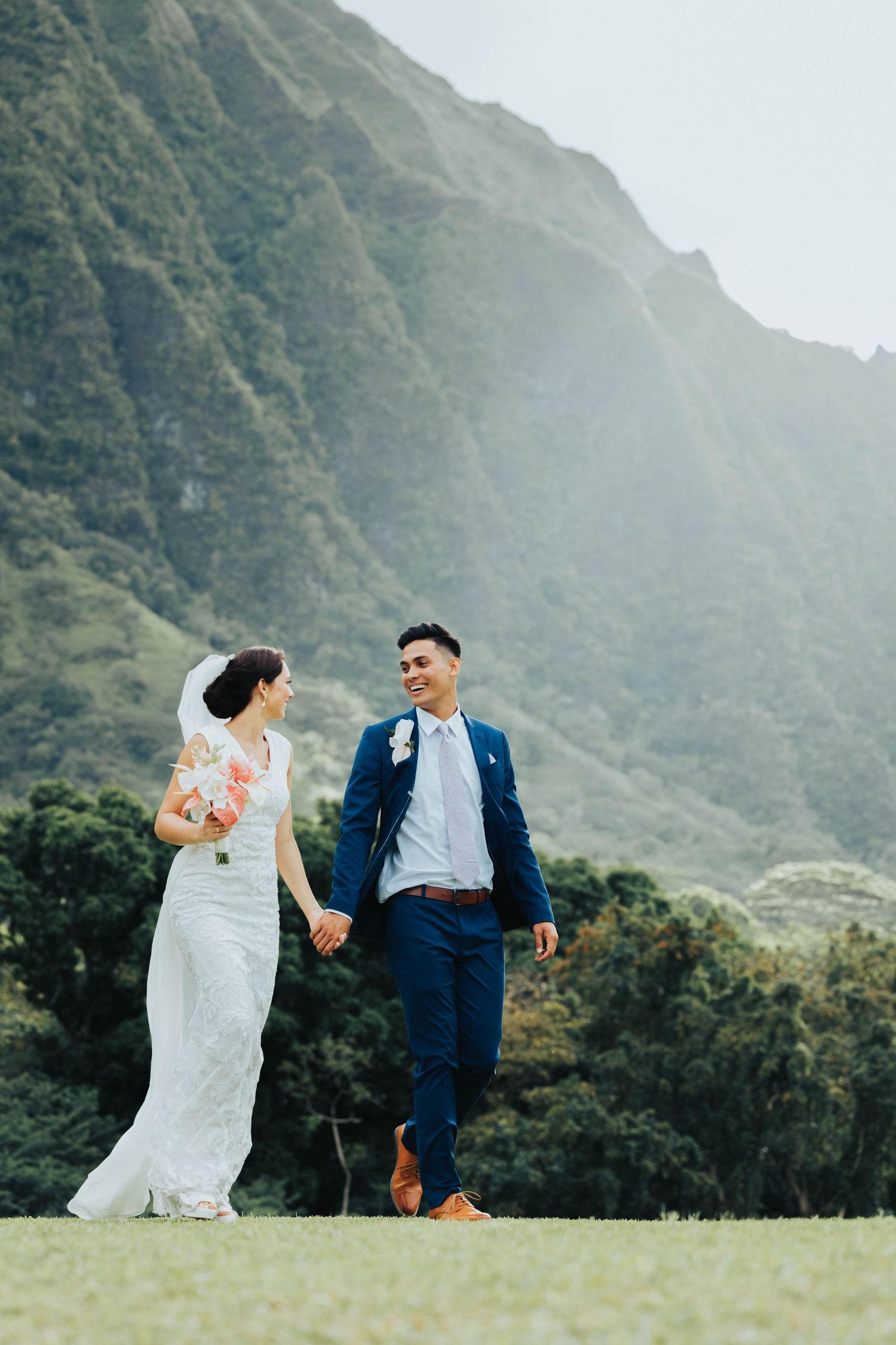 Couple in wedding attire shares playful moments against dramatic mountain backdrop in Hawaii's lush landscape.