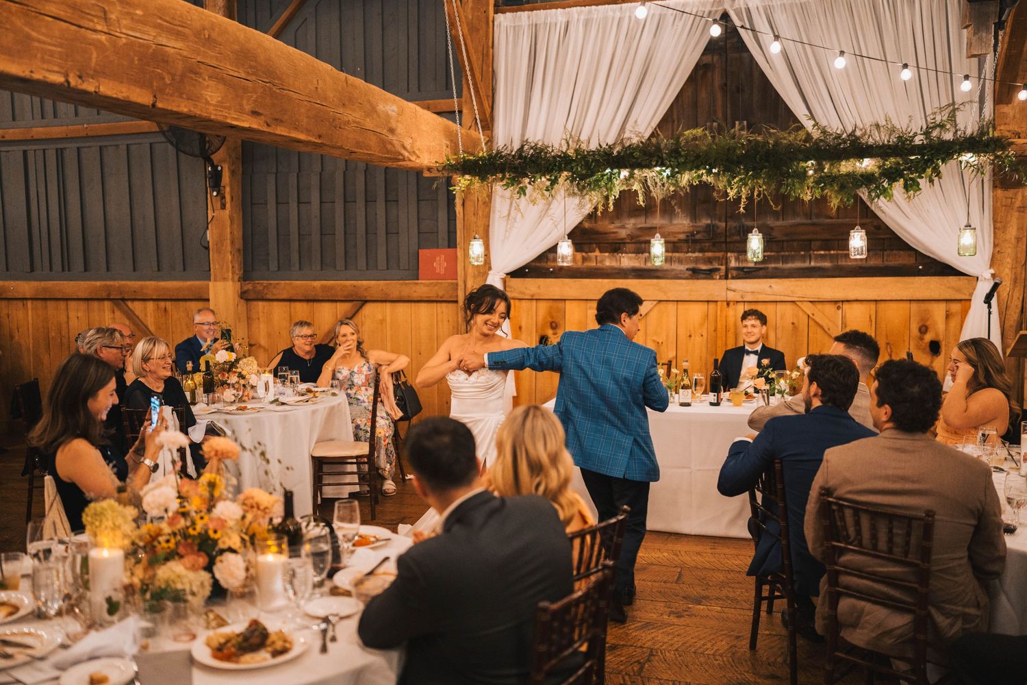 Rustic barn wedding reception with wooden beams and white drapes decorated with string lights and floral arrangements.