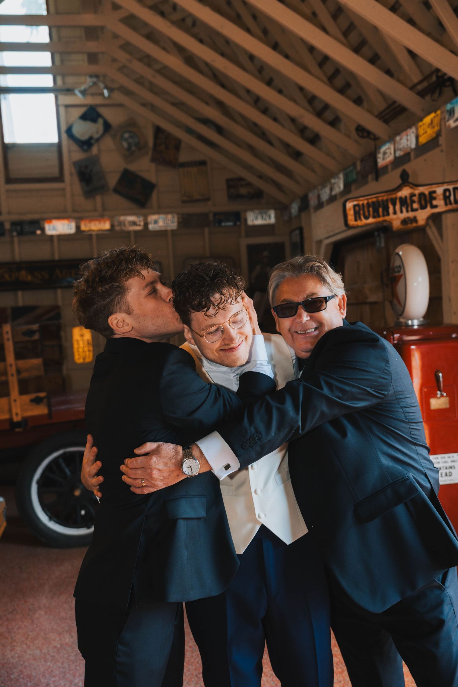 Three people in navy blue suits embrace and laugh together inside a rustic wooden building with exposed rafters.