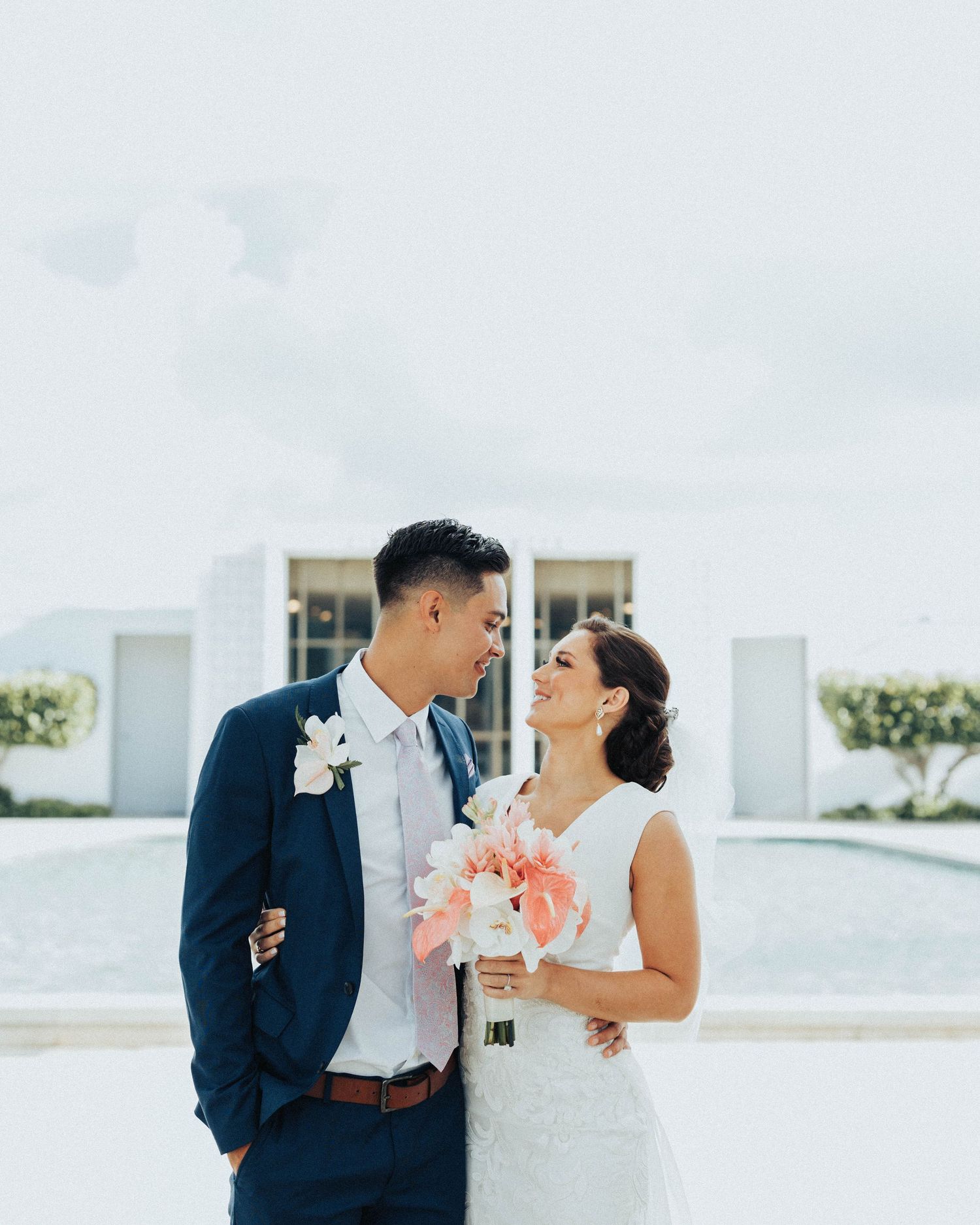 Newlywed couple in formal attire share an intimate moment while holding a coral-colored bouquet outdoors.