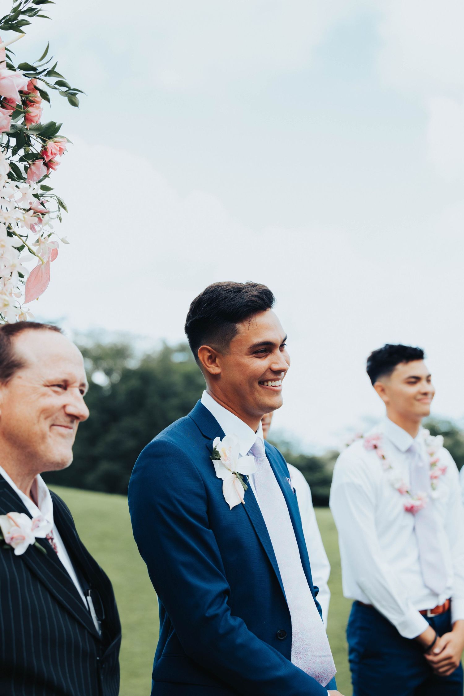Wedding party members in suits share a joyful moment outdoors with floral decorations visible in the background.