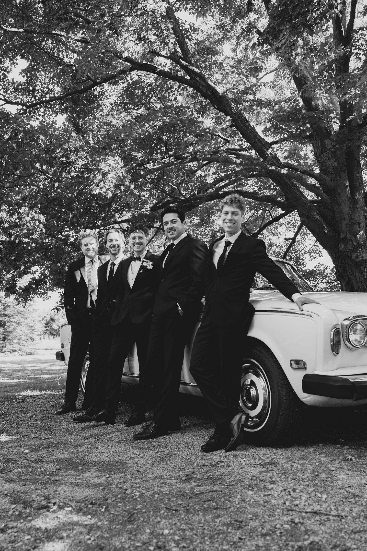 A group in formal black suits stands beside a classic white car under trees in a scenic outdoor setting.
