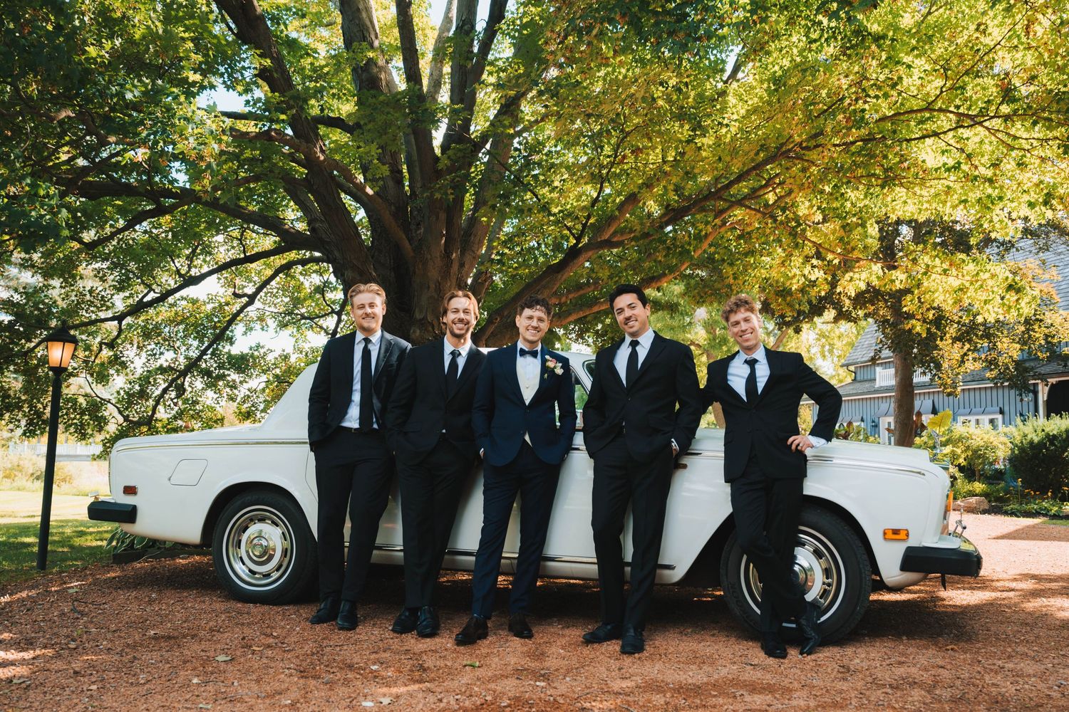 Groomsmen pose casually in front of a vintage car under fall trees with golden leaves.