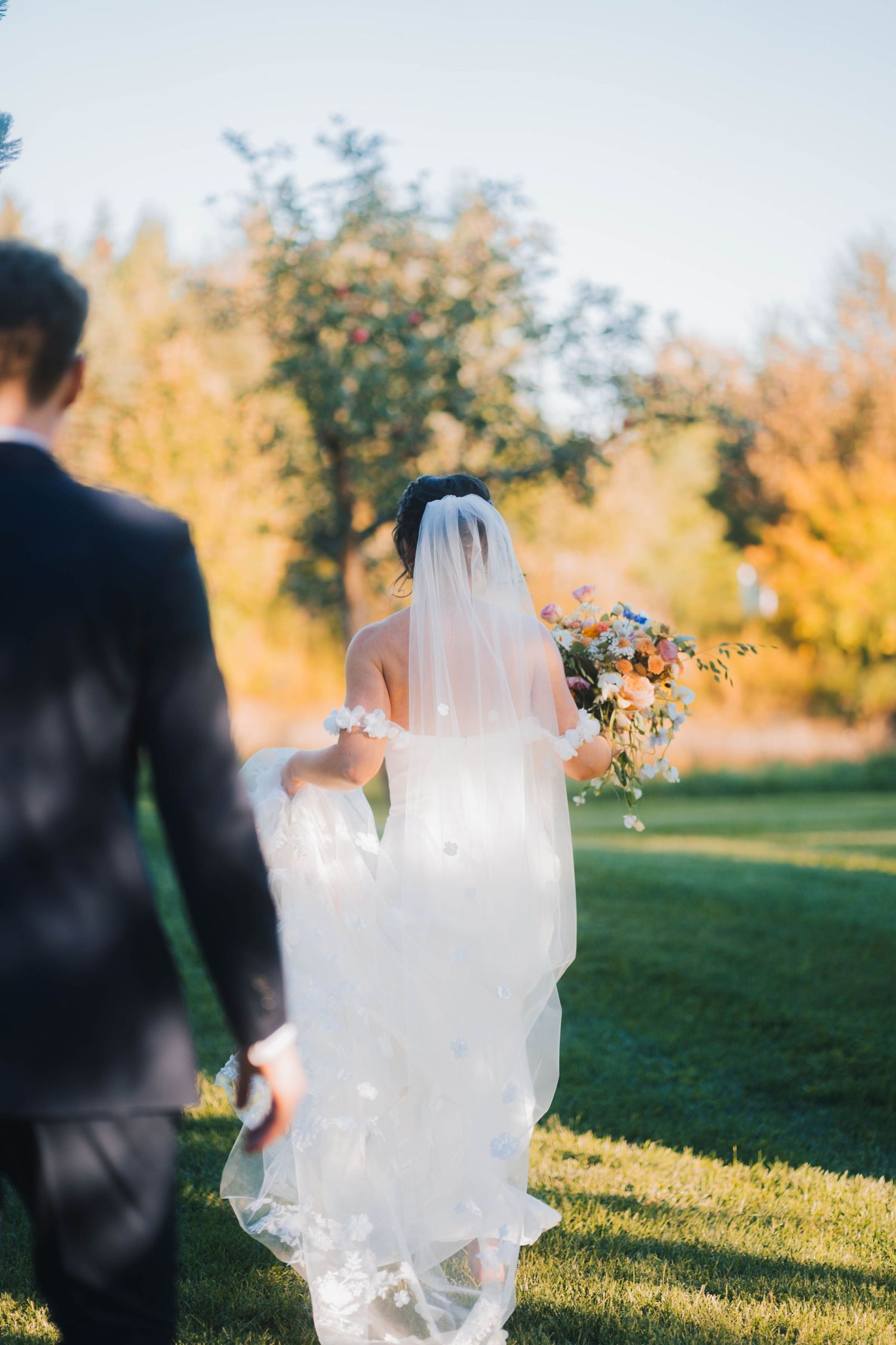 A couple in wedding attire stands in a sunlit meadow with golden autumn foliage in the background during sunset.