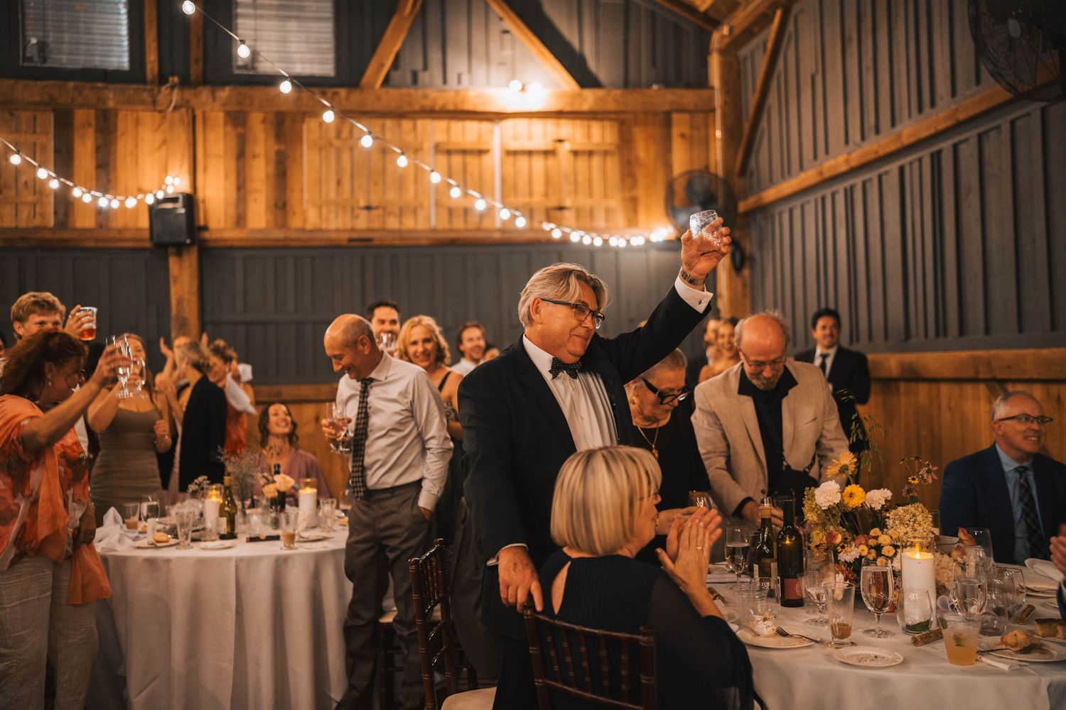Wedding reception guests seated at long tables in a rustic barn venue with warm lighting and elegant decor.