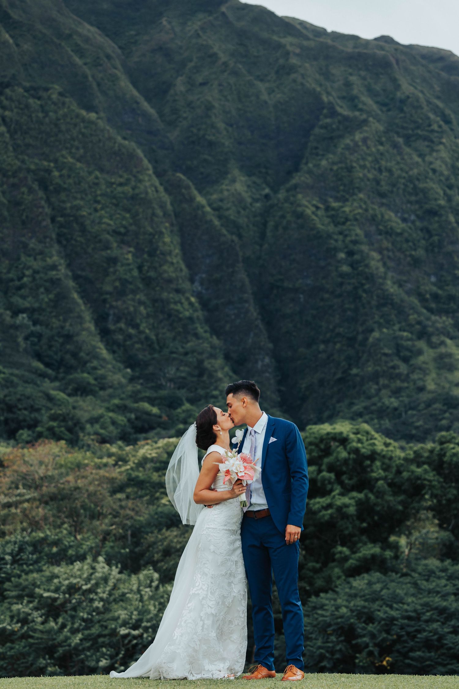 A couple in wedding attire embraces against a backdrop of lush green Hawaiian mountains on their special day.