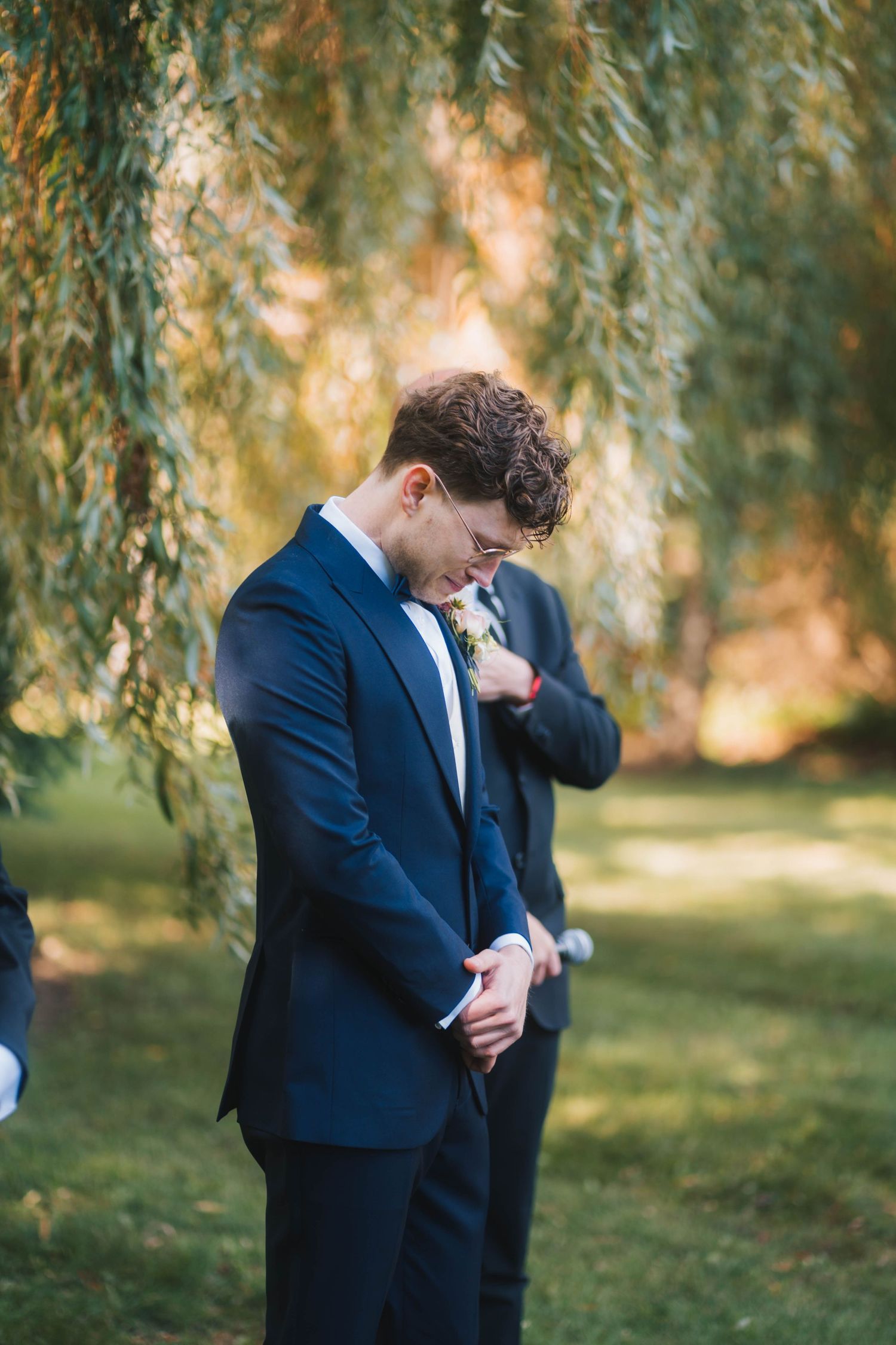 A person in a navy blue suit stands thoughtfully beneath drooping willow branches on a sunny day.