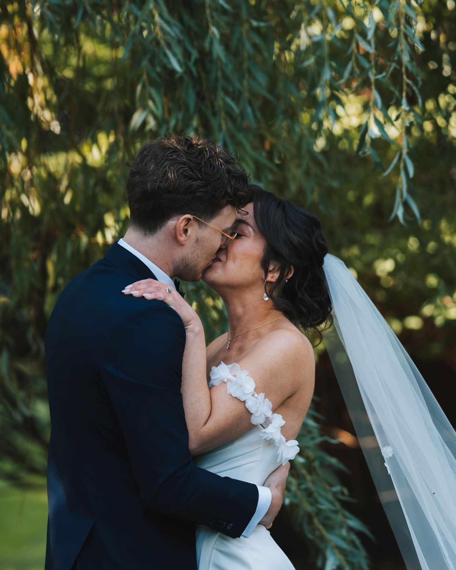 A romantic wedding couple shares a tender kiss under weeping willow trees in a garden setting.