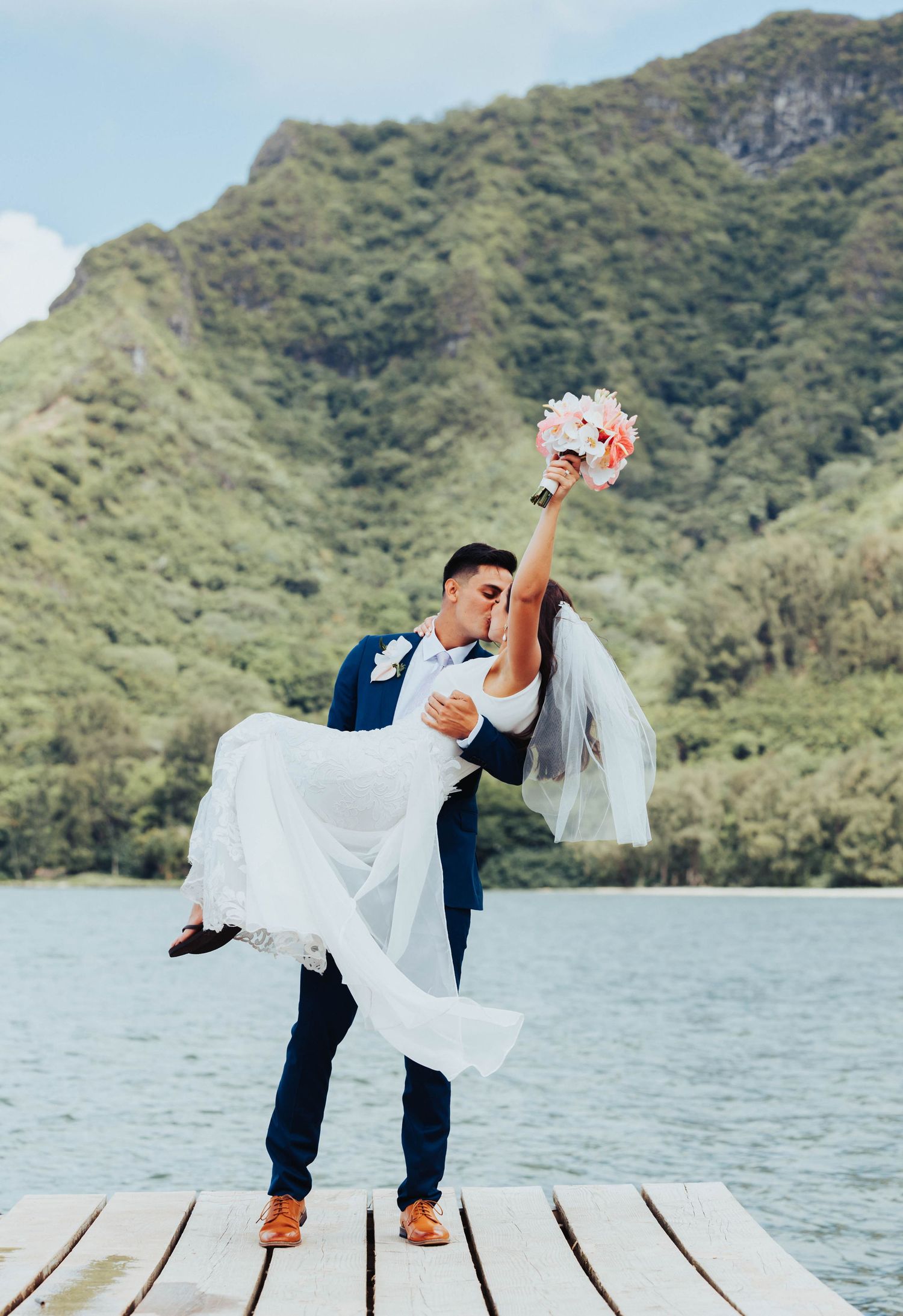 A couple in wedding attire share a romantic dip pose on a wooden dock with lush mountains and water in the background.