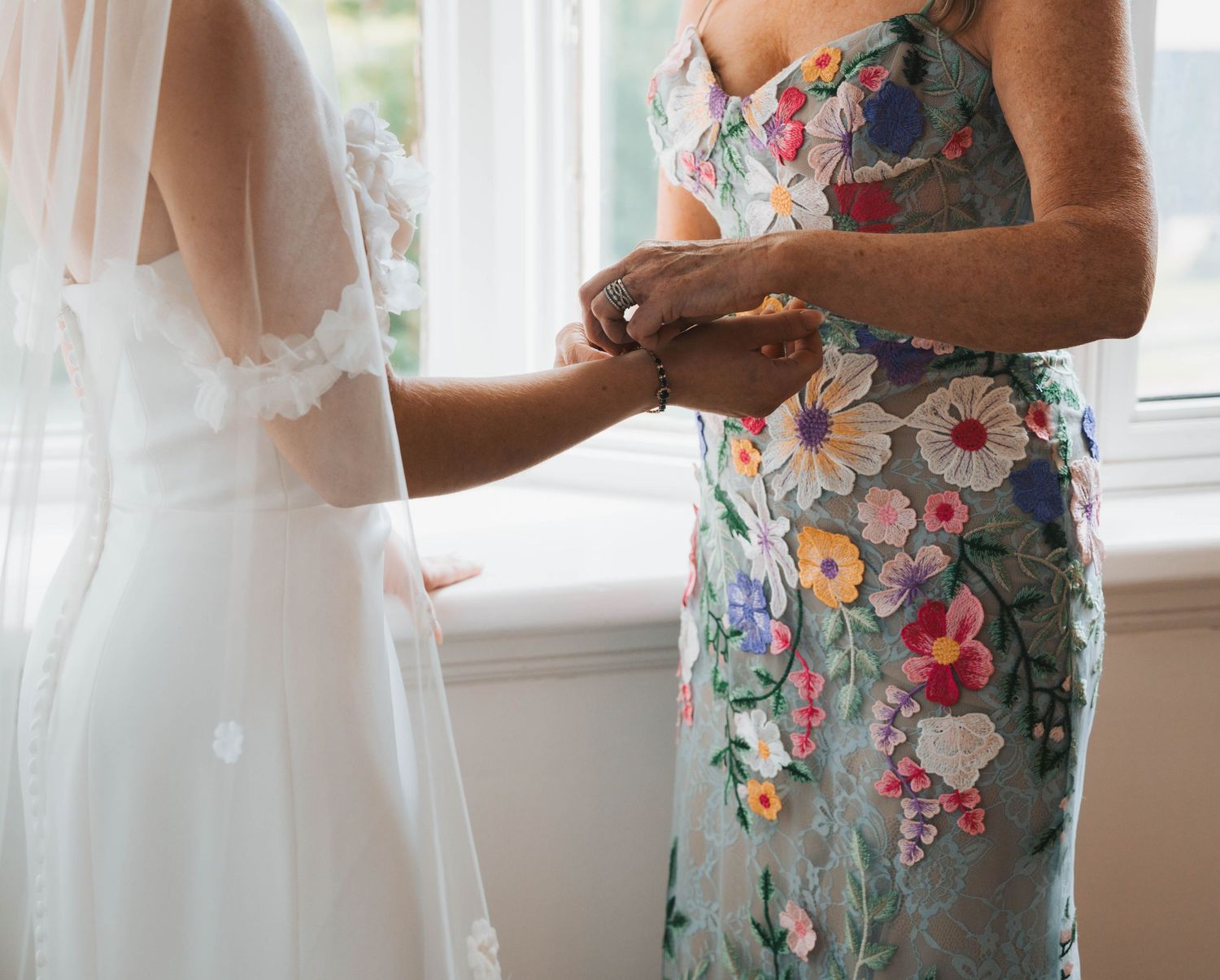 A person in a floral dress adjusts a white wedding gown hanging by a sunlit window.
