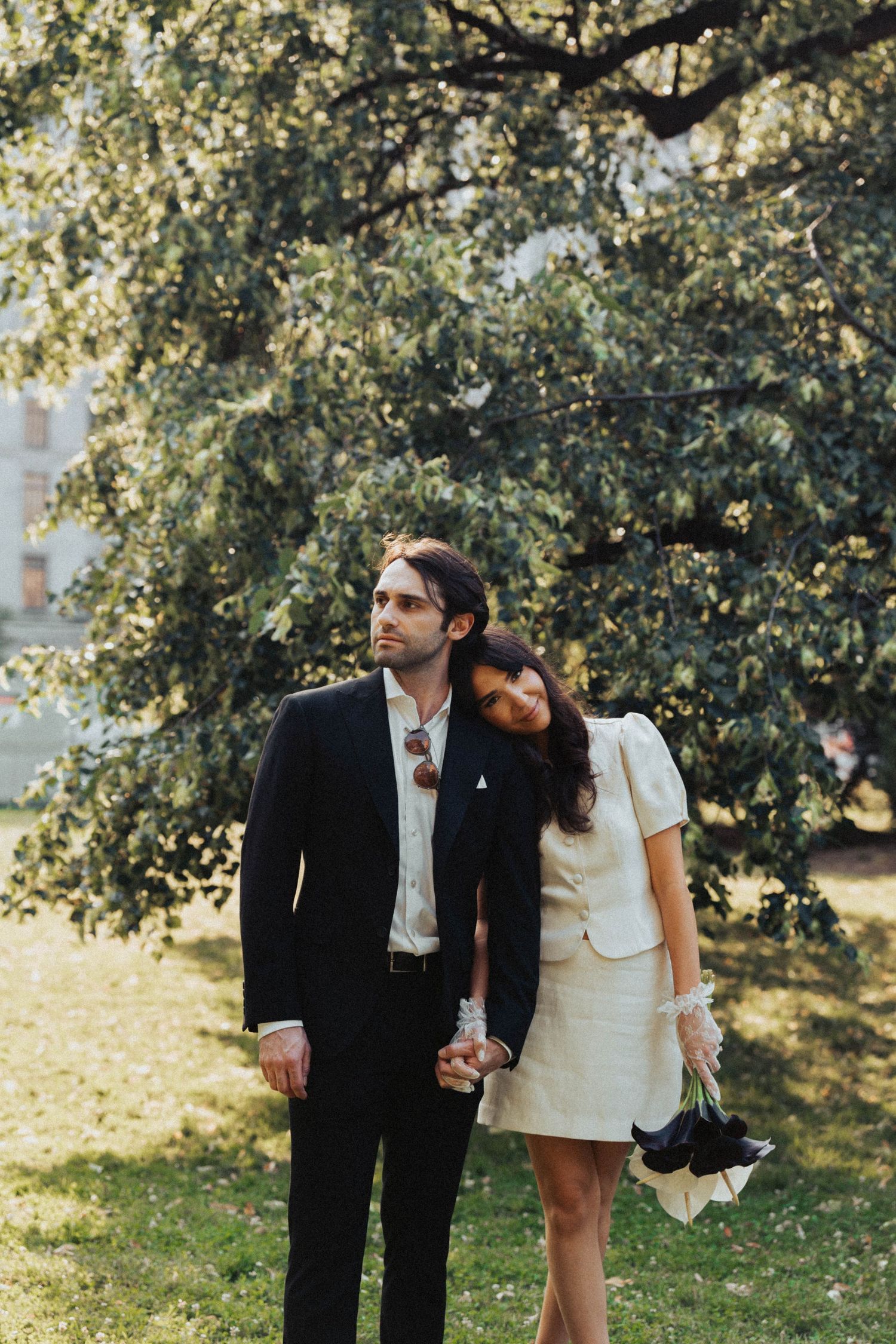 A romantic couple in formal attire shares tender moments under a sunlit tree in a park setting.