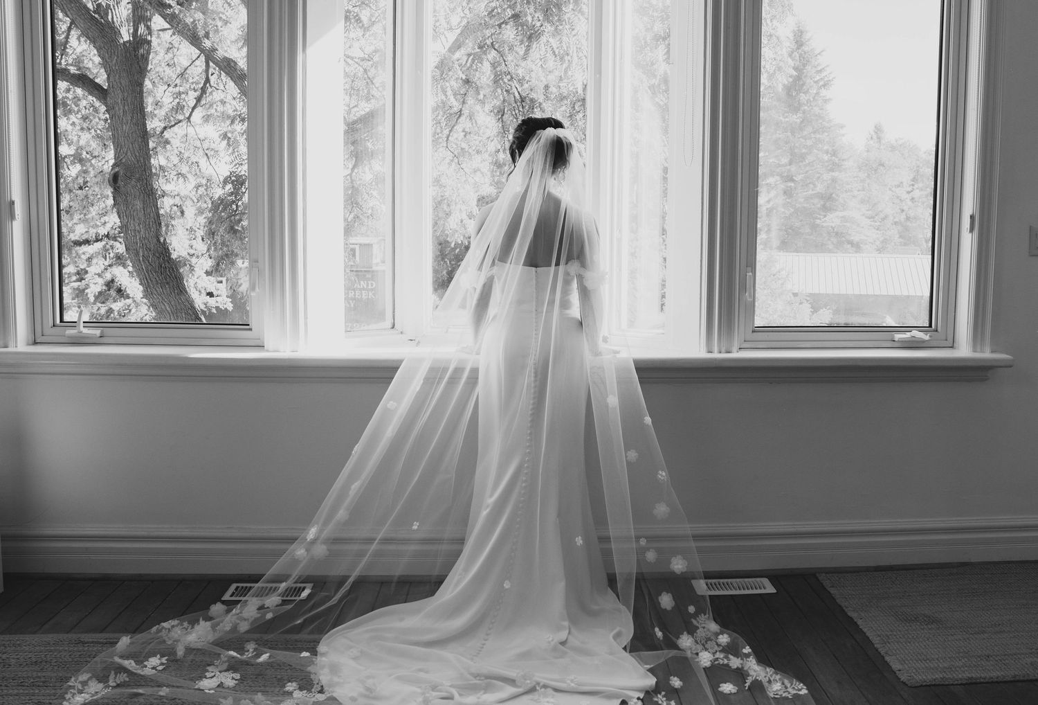A bride in a flowing white gown and veil stands elegantly by a window in black and white photography.