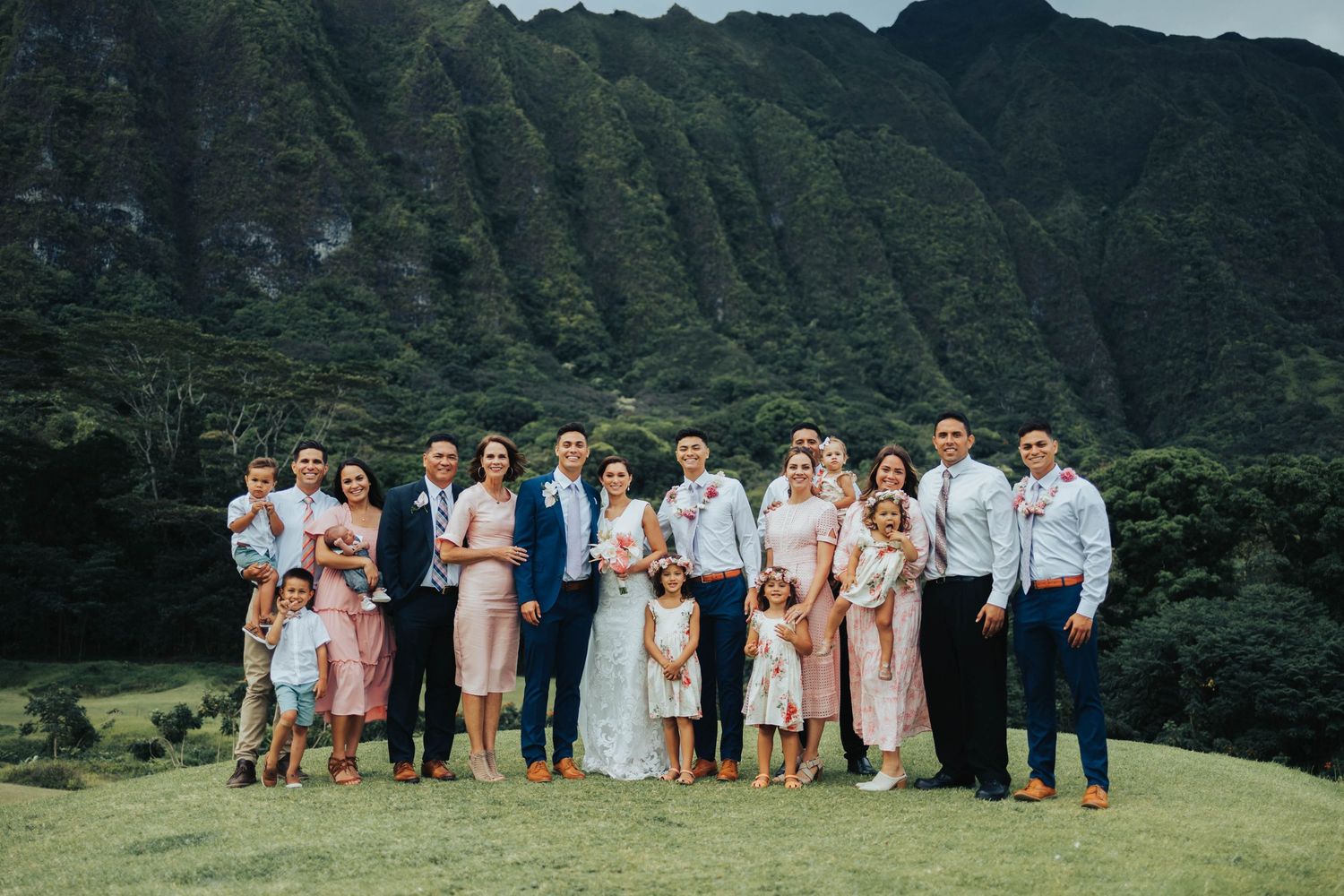 Wedding party stands together in front of lush green Hawaiian mountains on a sunny day.
