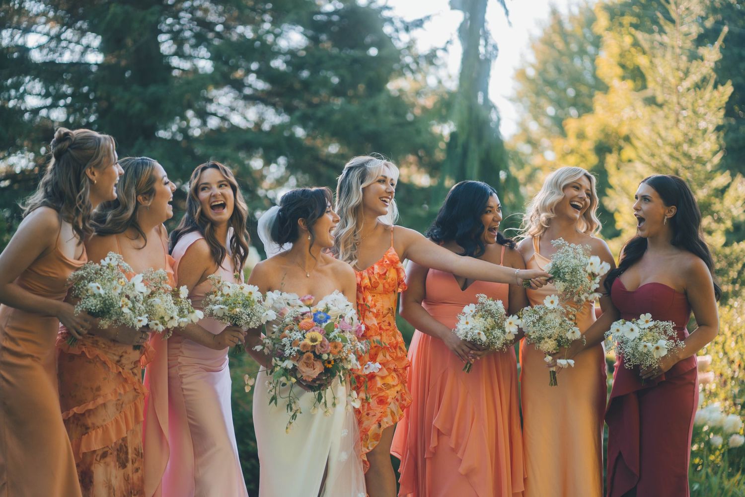Wedding party wearing colorful orange and burgundy dresses poses together outdoors during autumn celebration.