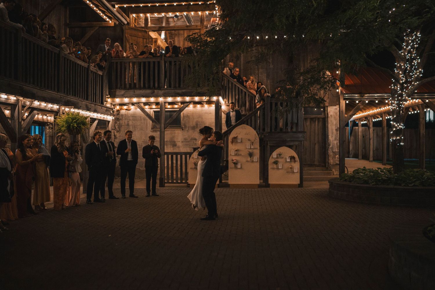 A string of warm glowing lights illuminates a rustic outdoor venue at dusk creating a romantic atmosphere.