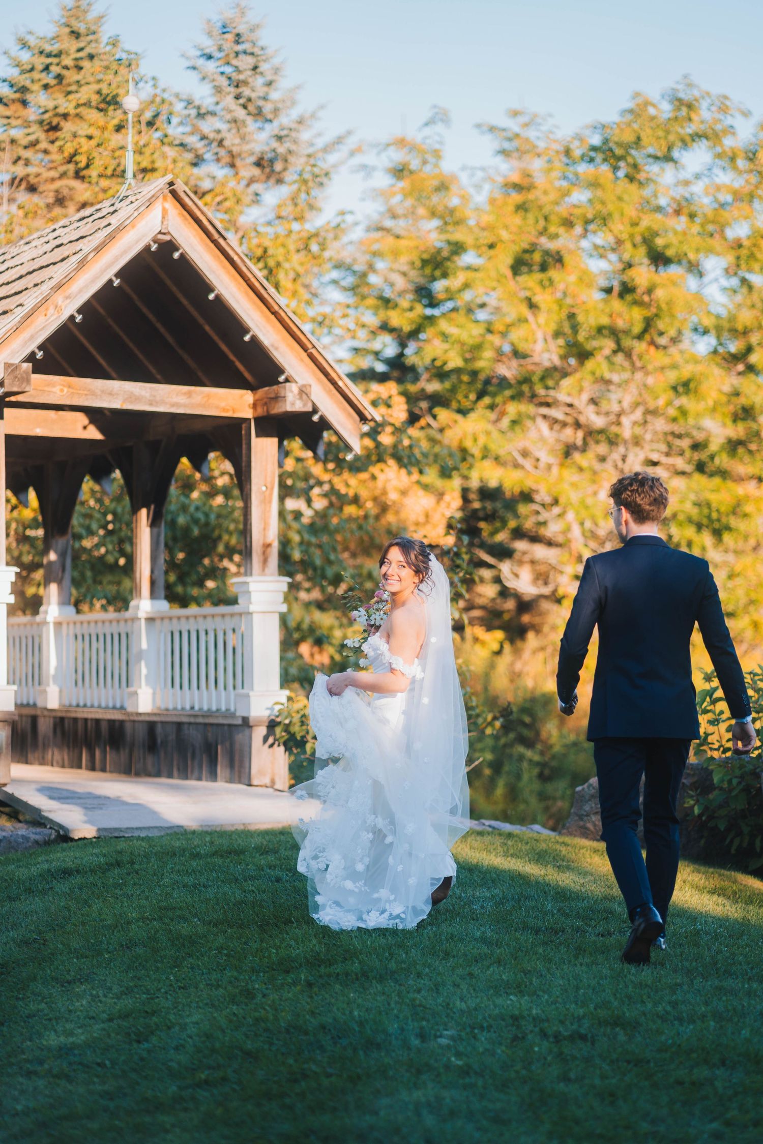 A couple in wedding attire walks across a grassy hill near a wooden gazebo during golden hour.