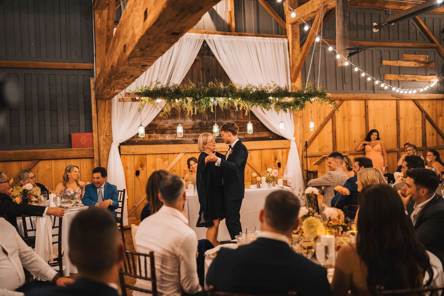 Wedding reception guests celebrate inside a rustic wooden barn decorated with string lights and white drapery.
