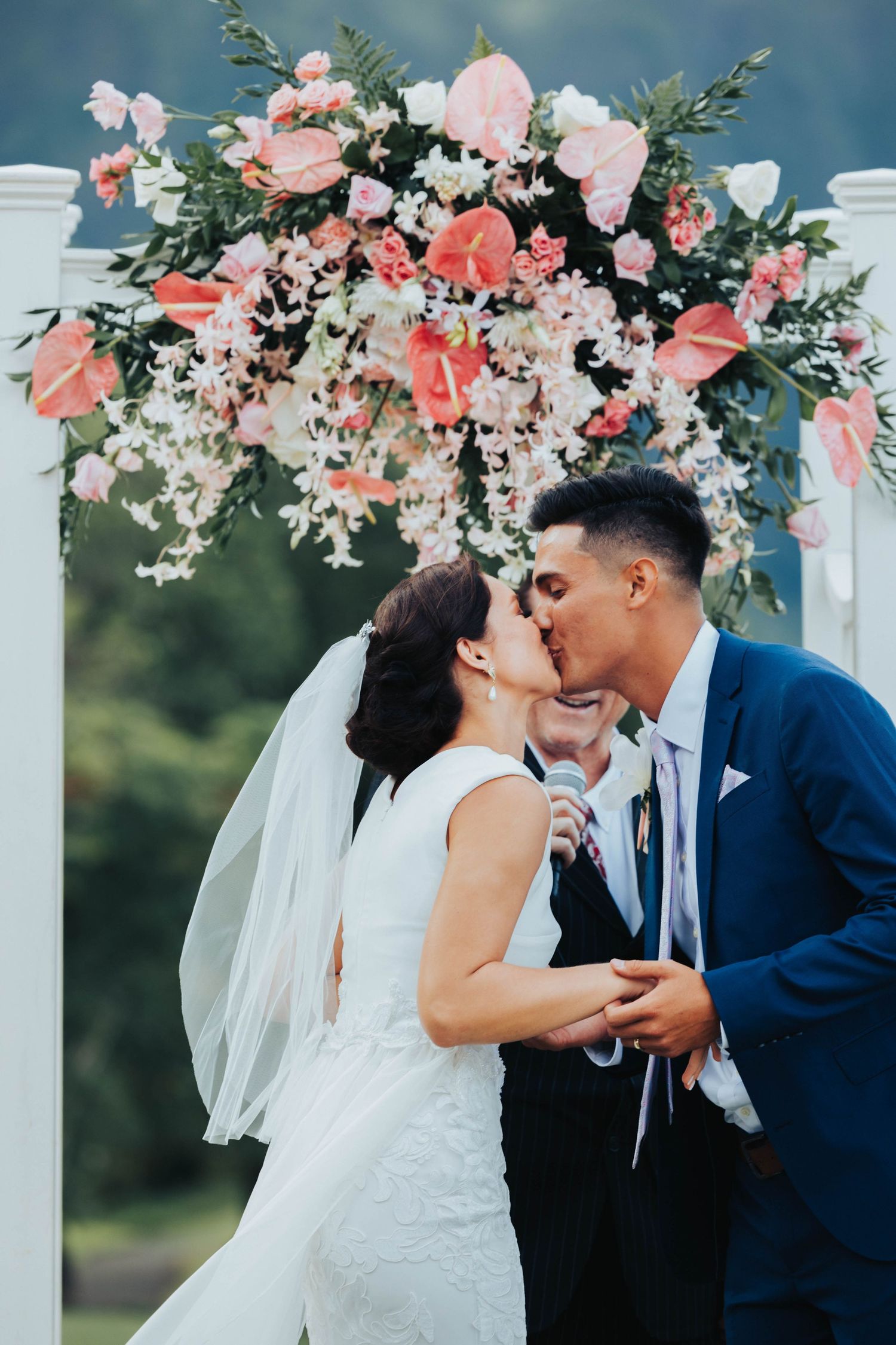 Newlyweds share a romantic kiss under an elegant floral arch decorated with pink and white blooms at an outdoor ceremony.