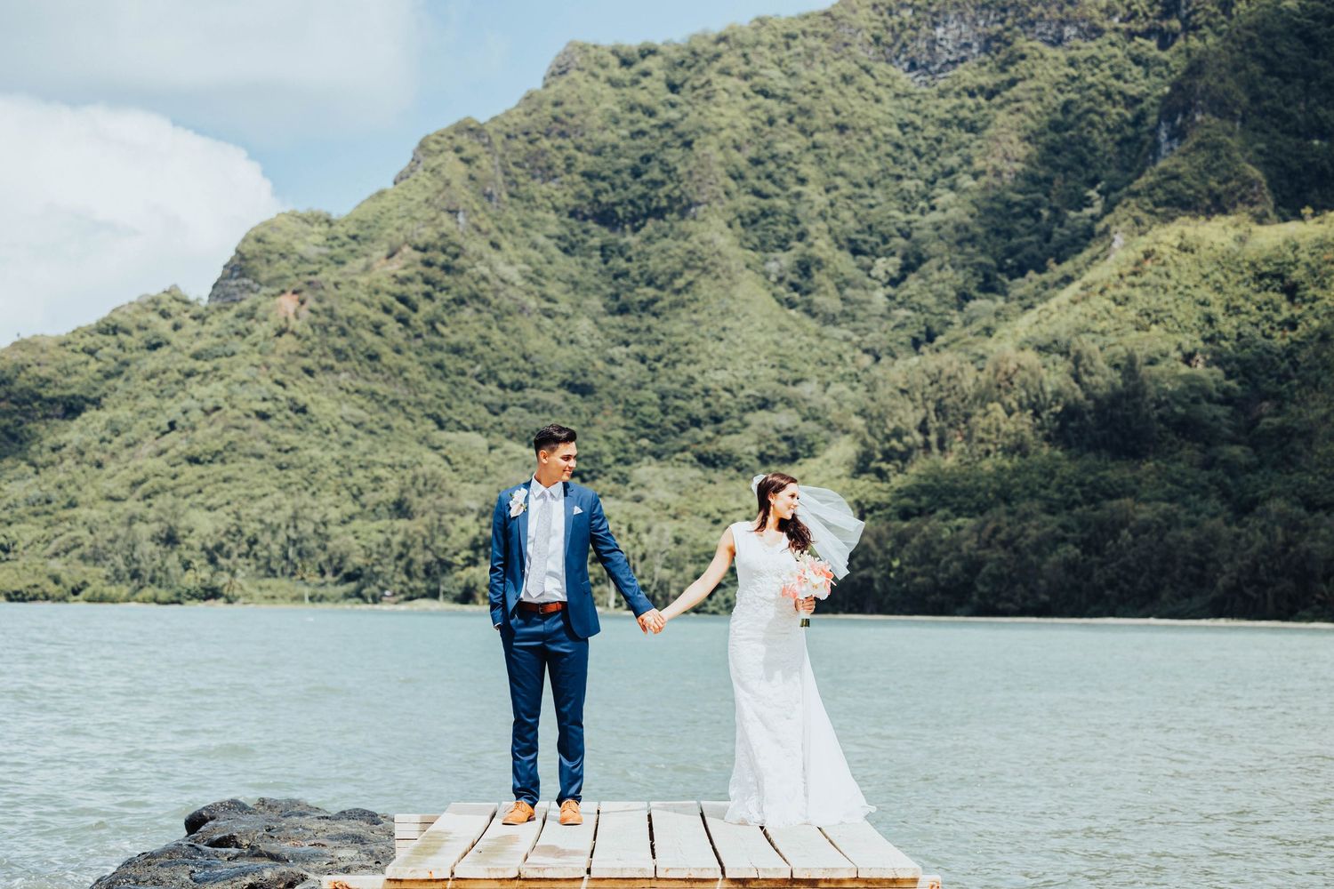 A couple in wedding attire walks along a scenic mountain lake shoreline on their special day.