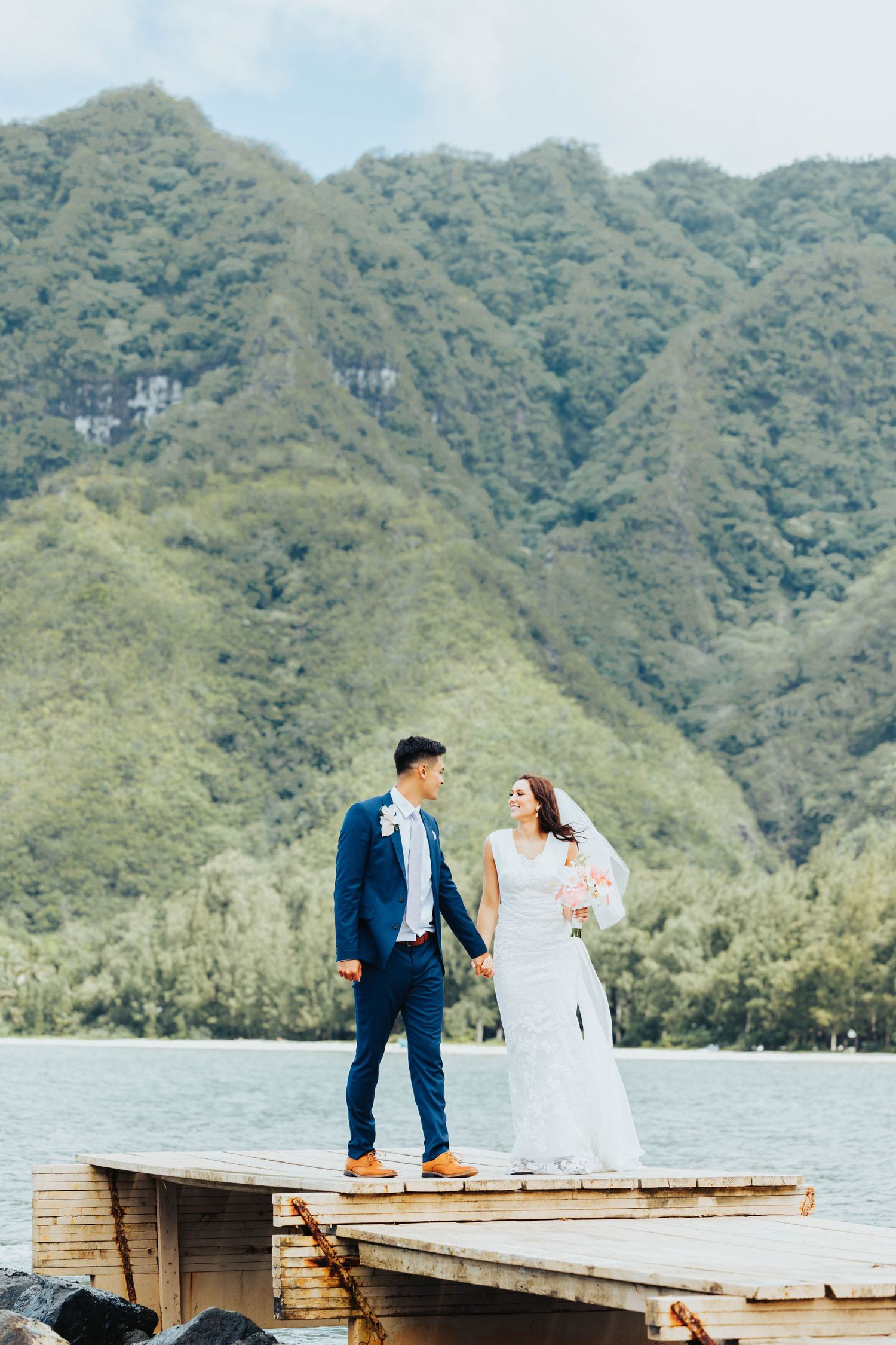 A couple in wedding attire stands on a wooden dock overlooking a scenic mountain lake landscape.