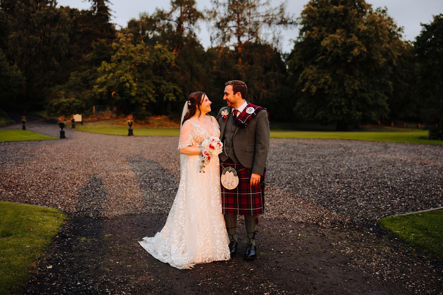 Bride and groom sharing a romantic moment at golden hour, groom in red tartan kilt on gravel path.