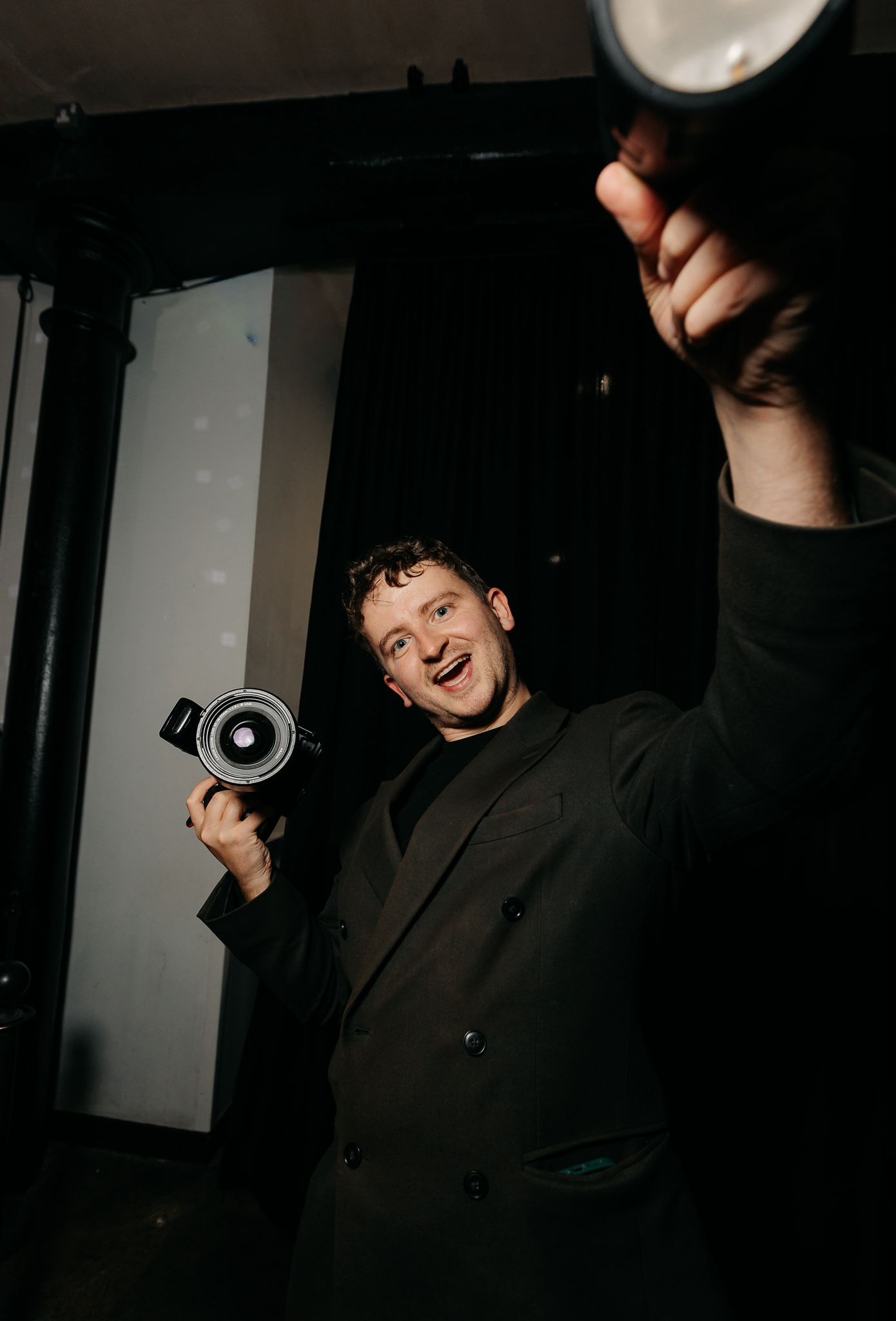 Smiling photographer in black suit holds camera while celebrating at a dark venue.