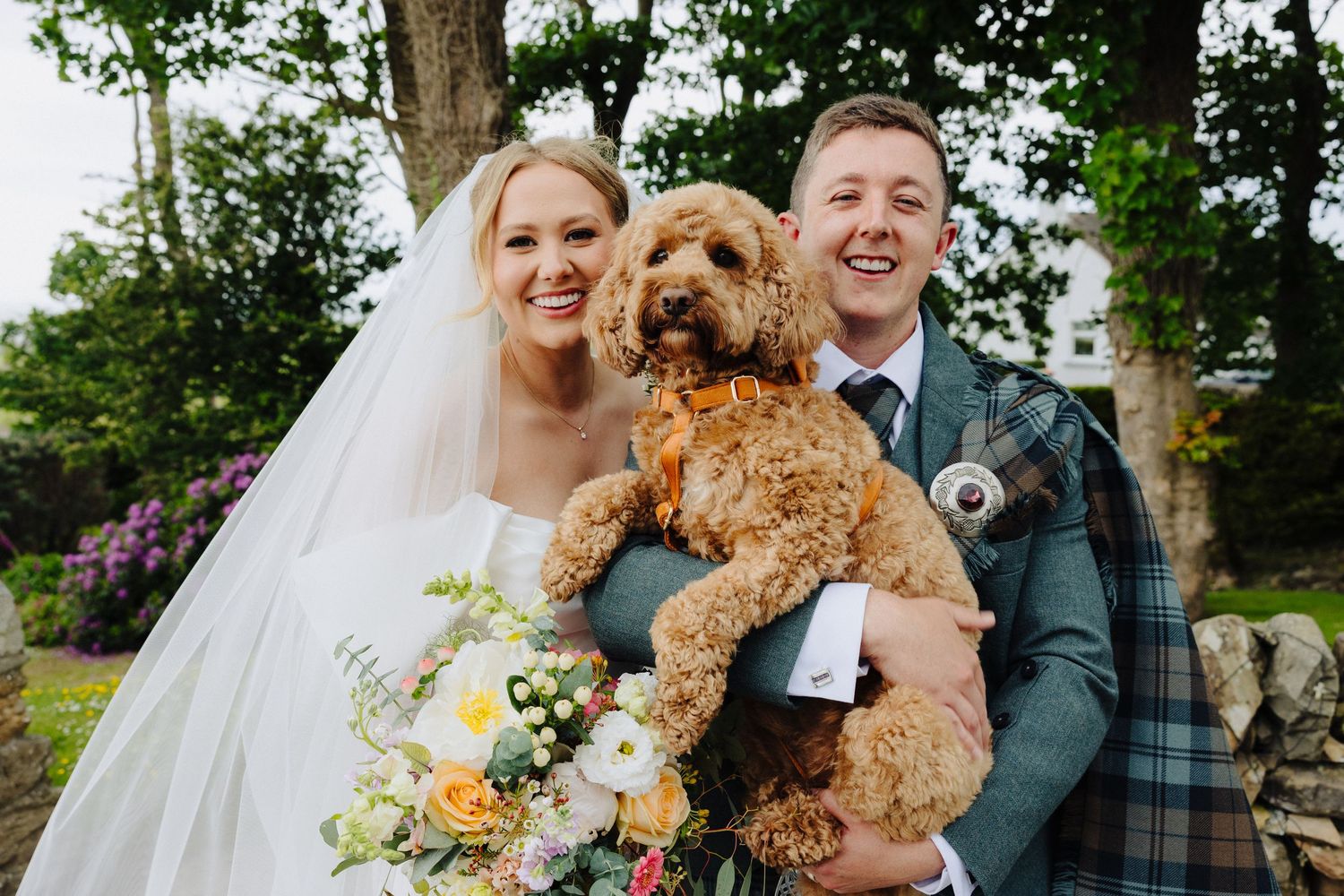 Smiling bride and groom in Scottish tartan kilt holding a fluffy golden cockapoo dog on their wedding day.