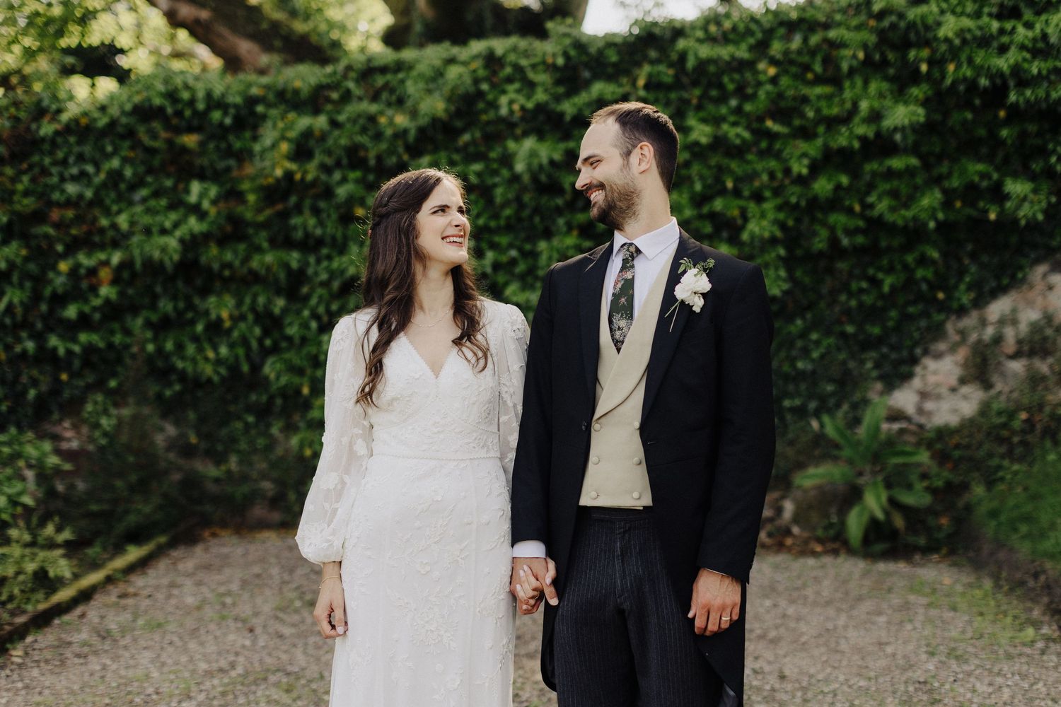 Bride in lace dress and groom in navy suit holding hands in lush green garden setting.