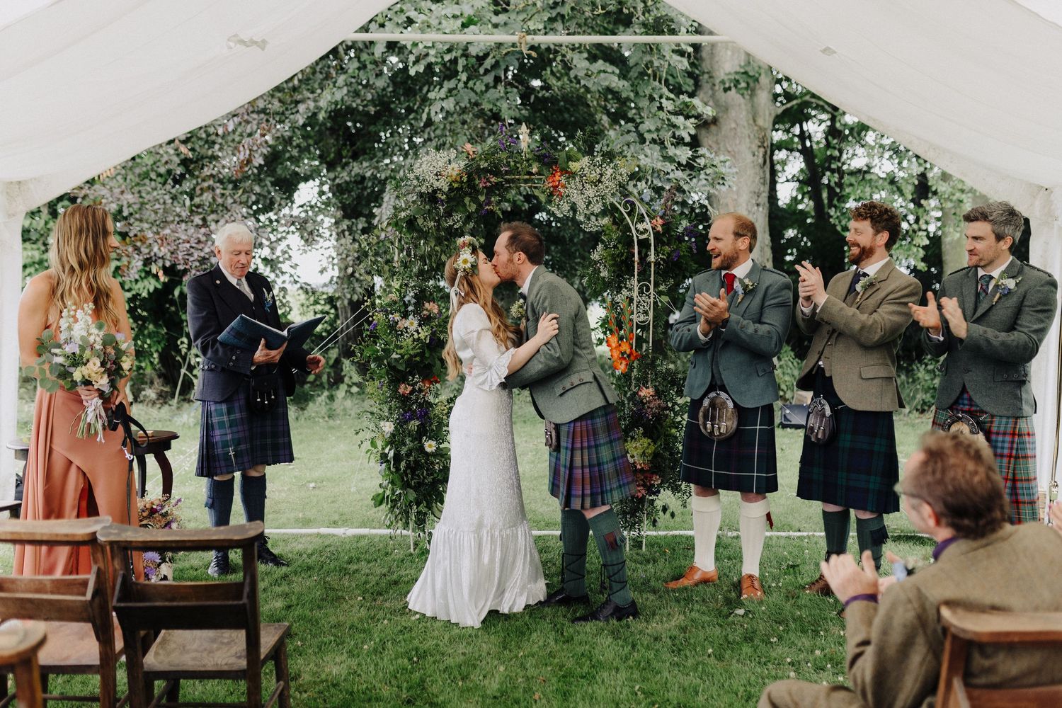Bride and groom sharing first kiss at outdoor tent ceremony surrounded by guests in tartan kilts.