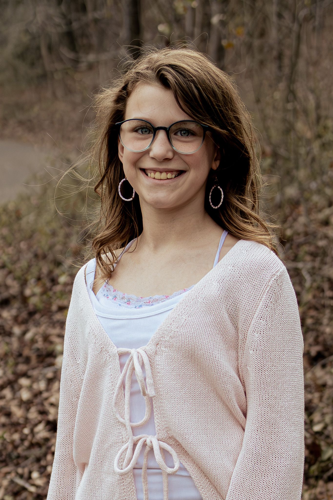 Smiling teenage girl with glasses and hoop earrings wearing a pink cardigan poses outdoors in a wooded setting.