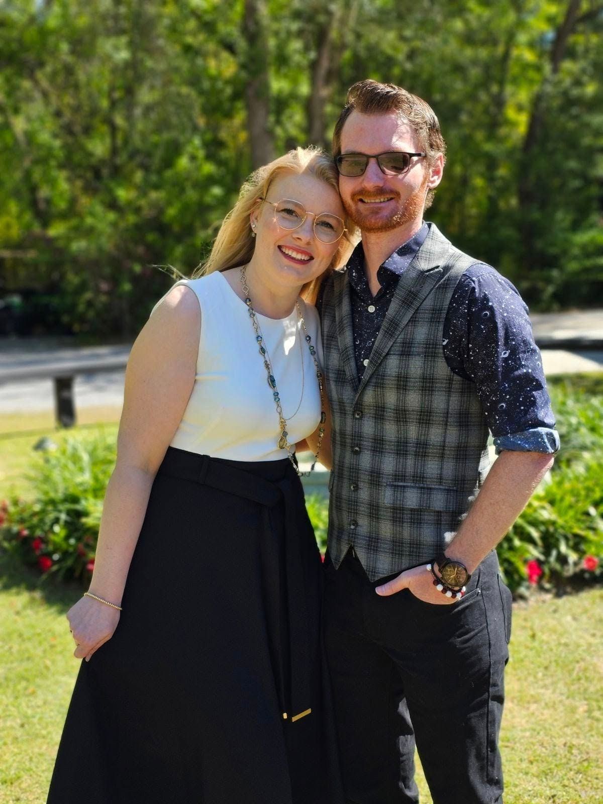 A smiling couple poses outdoors near lush green trees, woman in white top and black skirt, man in plaid vest.
