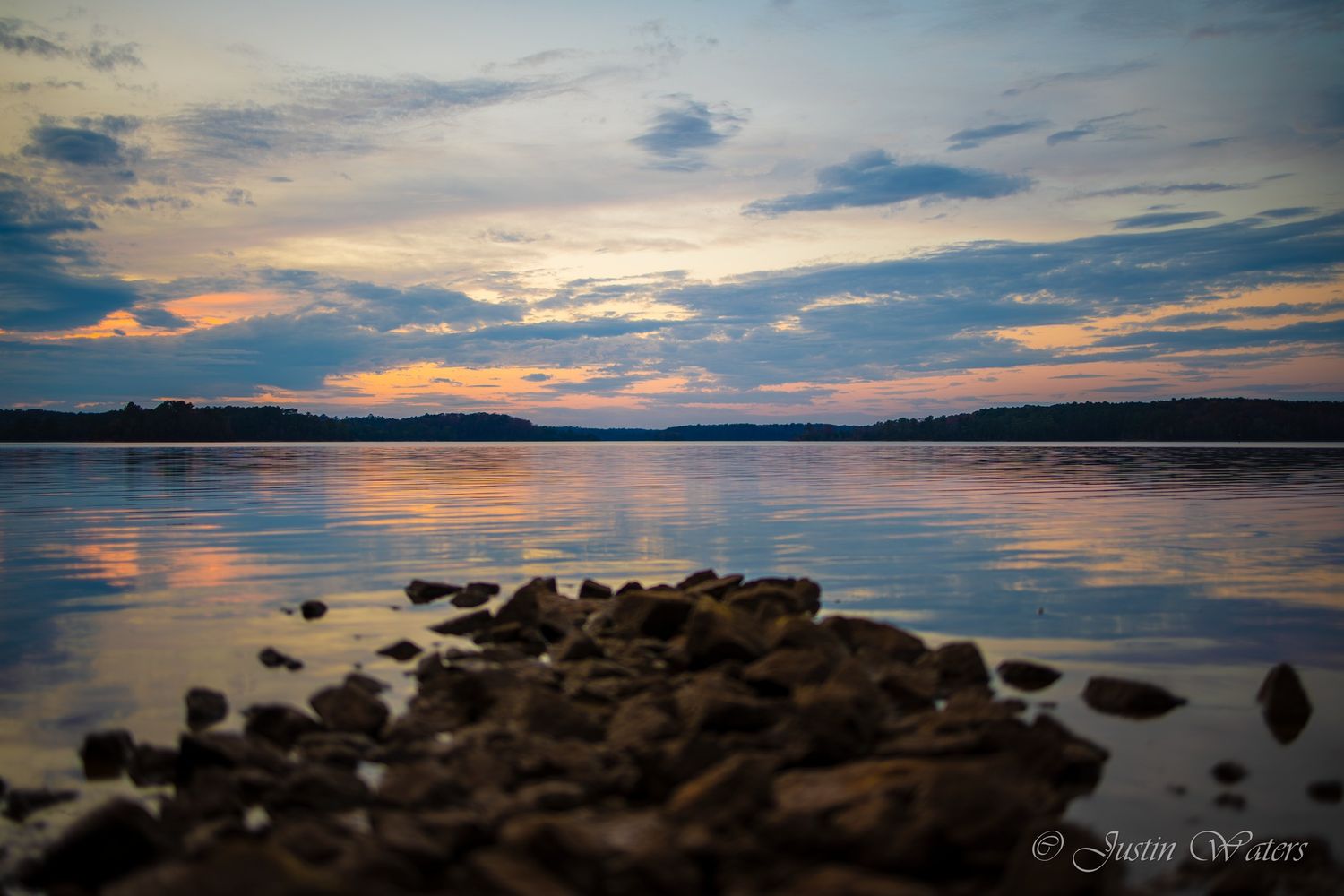 Serene sunset over calm lake with rocky shoreline reflecting colorful clouds in still water.