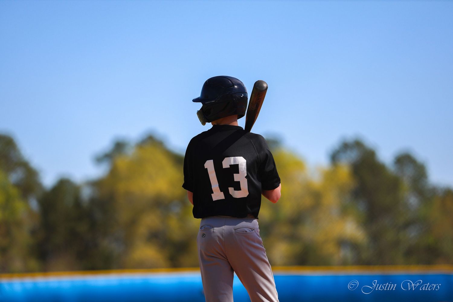 Youth baseball player wearing number 13 jersey stands at bat during a sunny outdoor game.