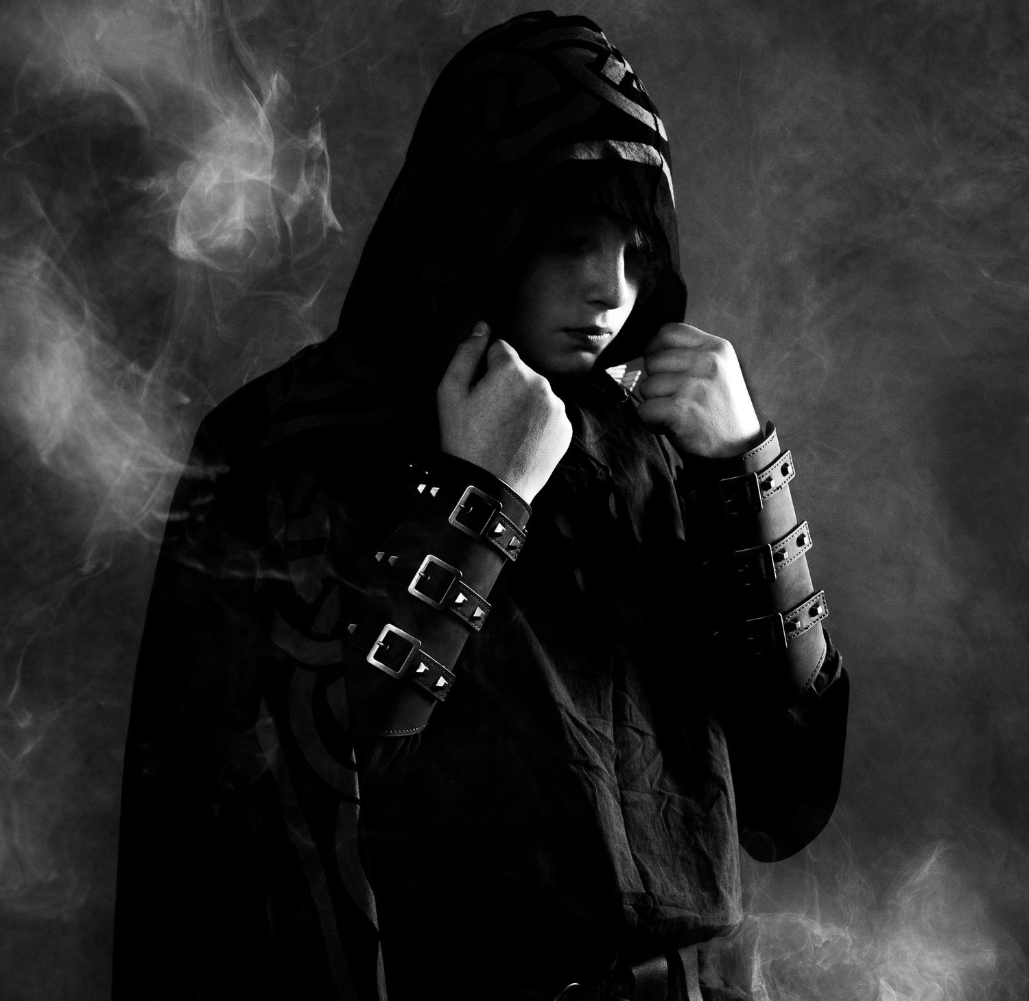 Dramatic black and white portrait of a person in dark hooded cloak with leather bracers surrounded by smoke.
