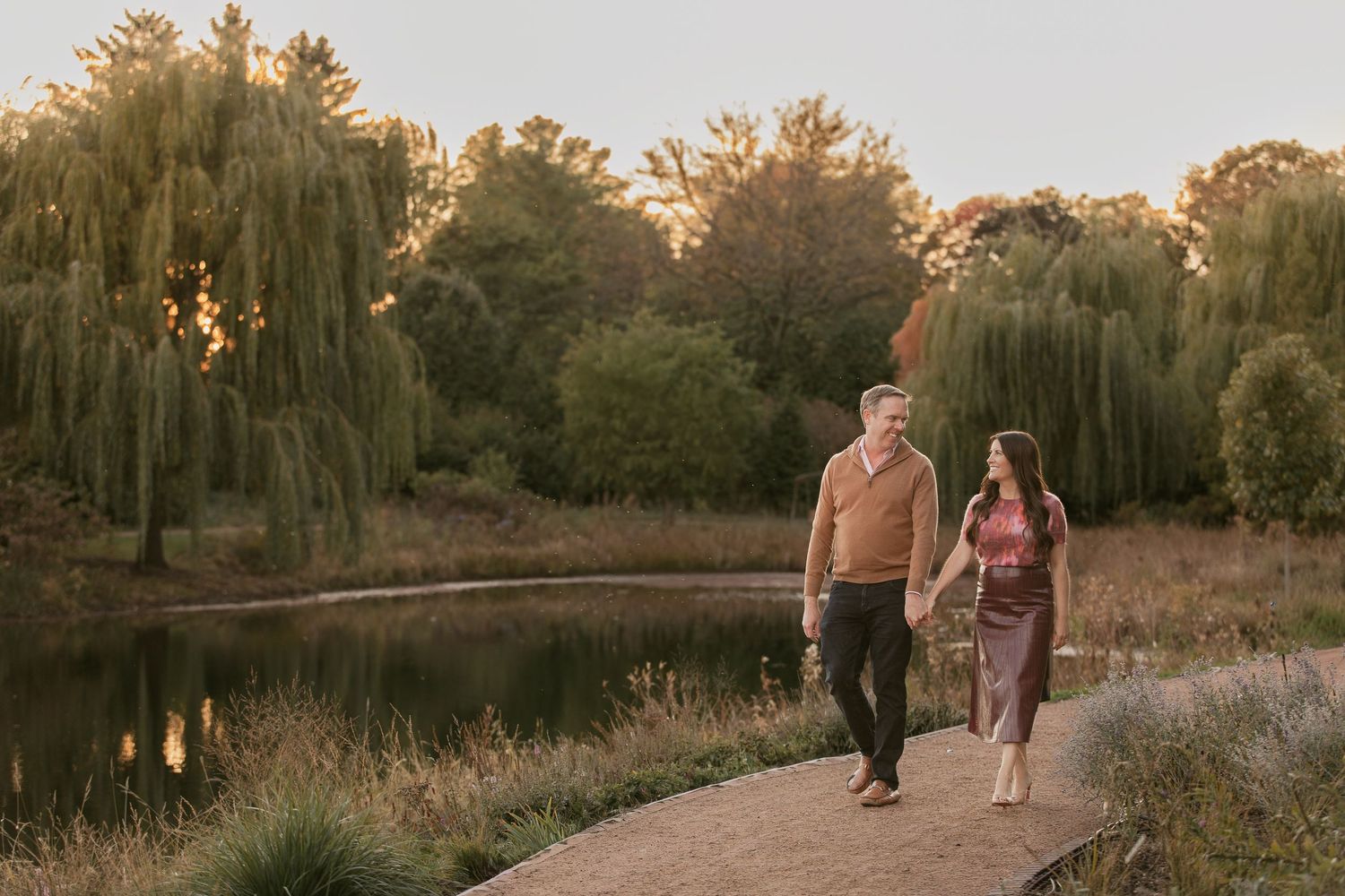 A couple walks along a winding path through an autumn park at sunset with golden light filtering through trees.