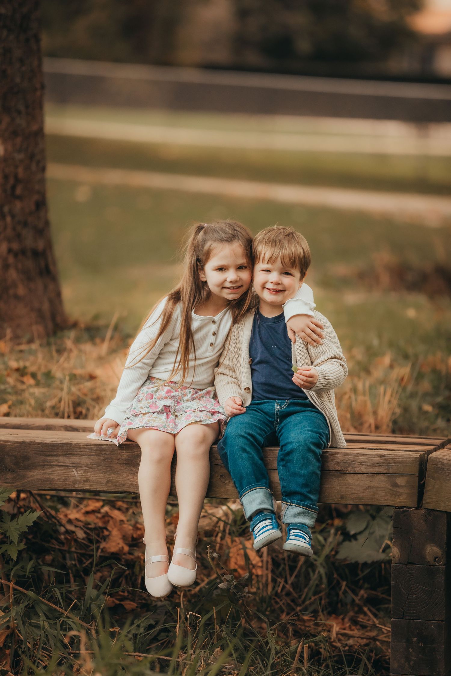 Two children sit together on a wooden bench in an outdoor autumn setting wearing casual clothes.