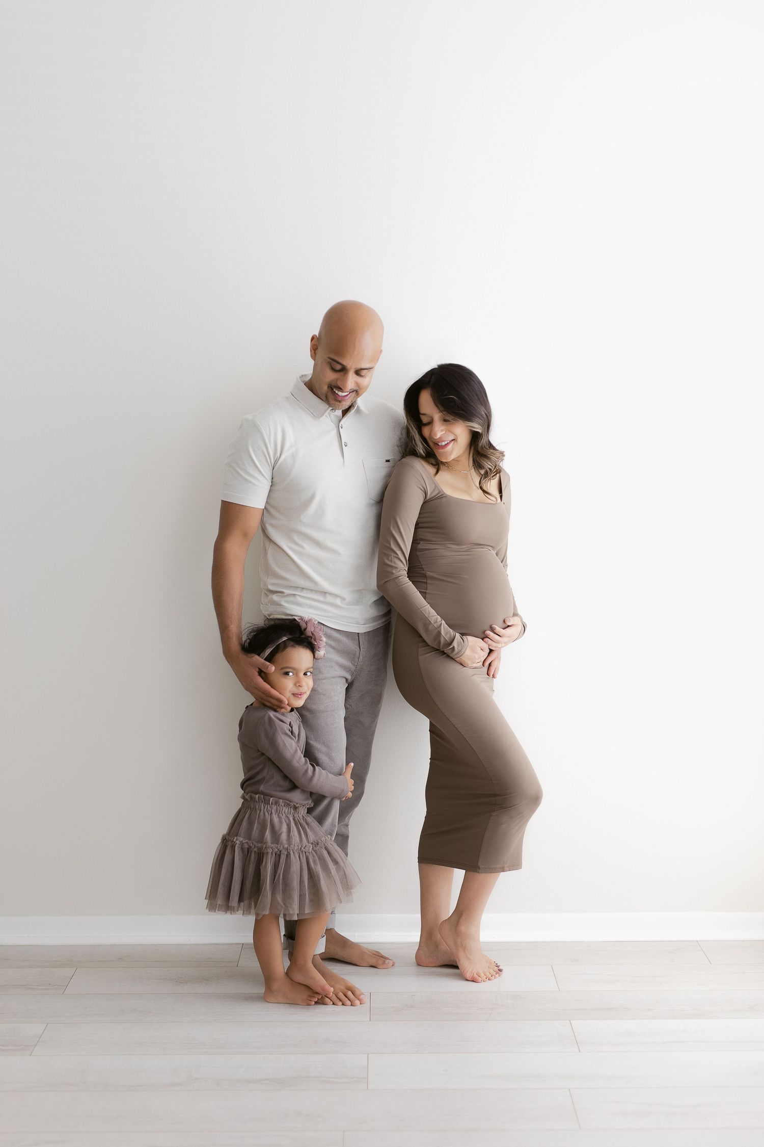 Minimalist maternity photo shows family of three posing against white wall in neutral tones.