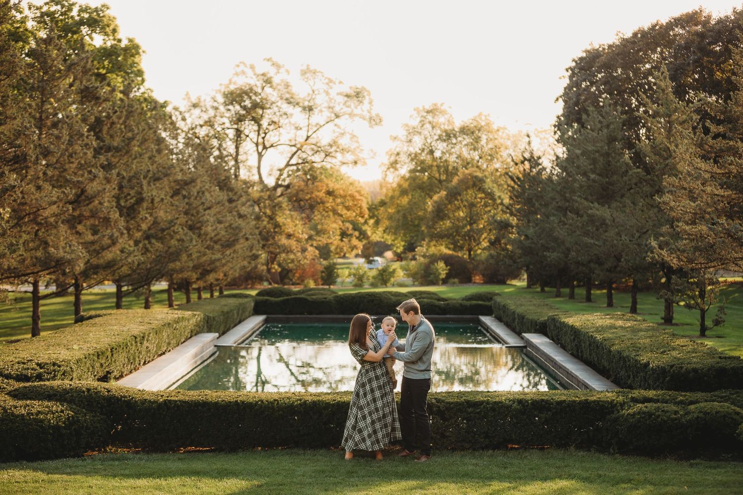 Couple embracing in a formal garden with manicured hedges and reflecting pool at golden hour.