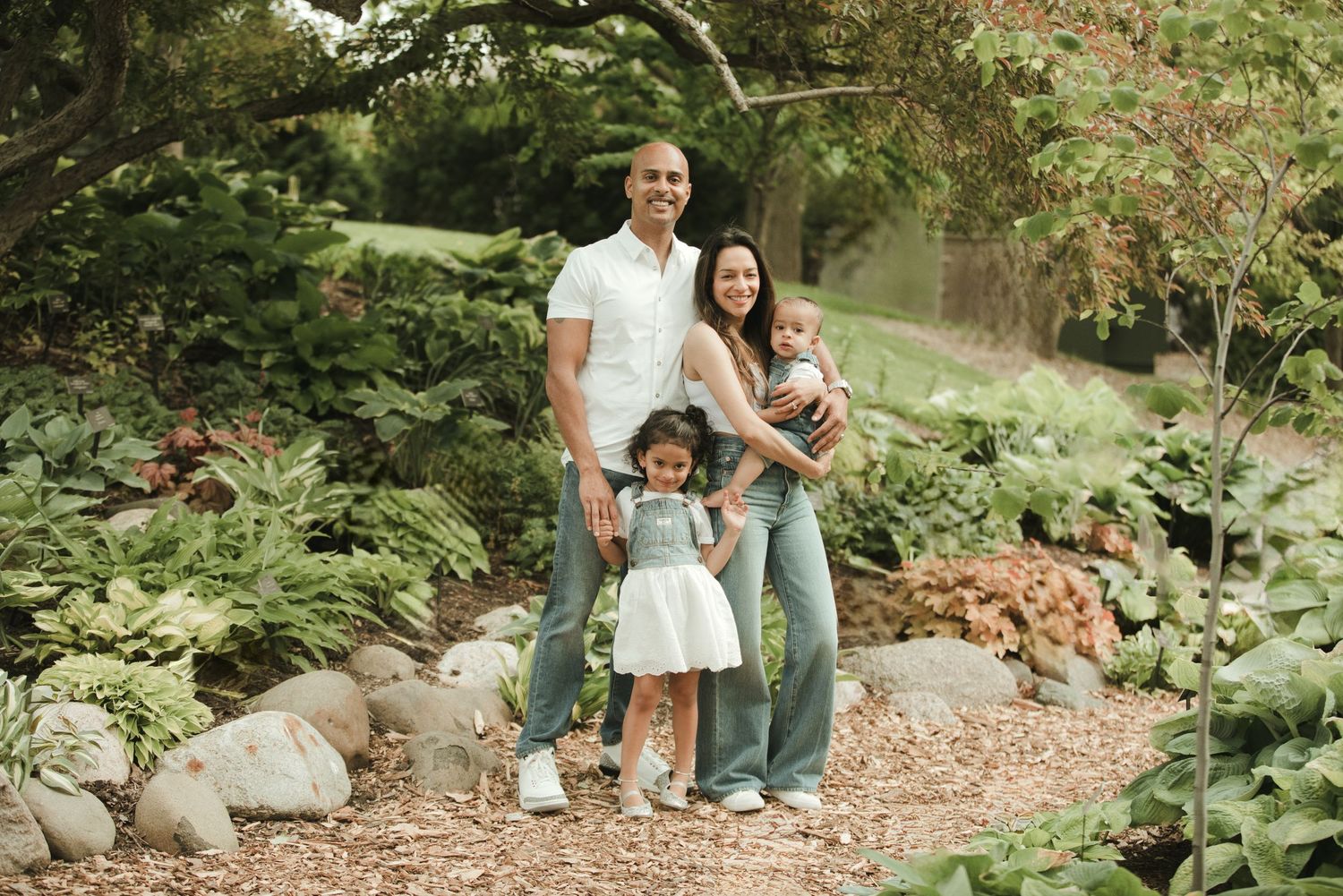 Happy family of four posing on a garden path surrounded by lush green plants and rocks.