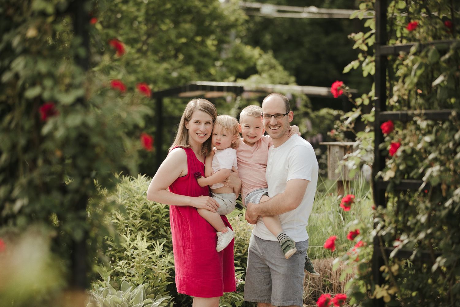 Happy family of four posing in a lush garden with red roses, parents holding two young children outdoors.
