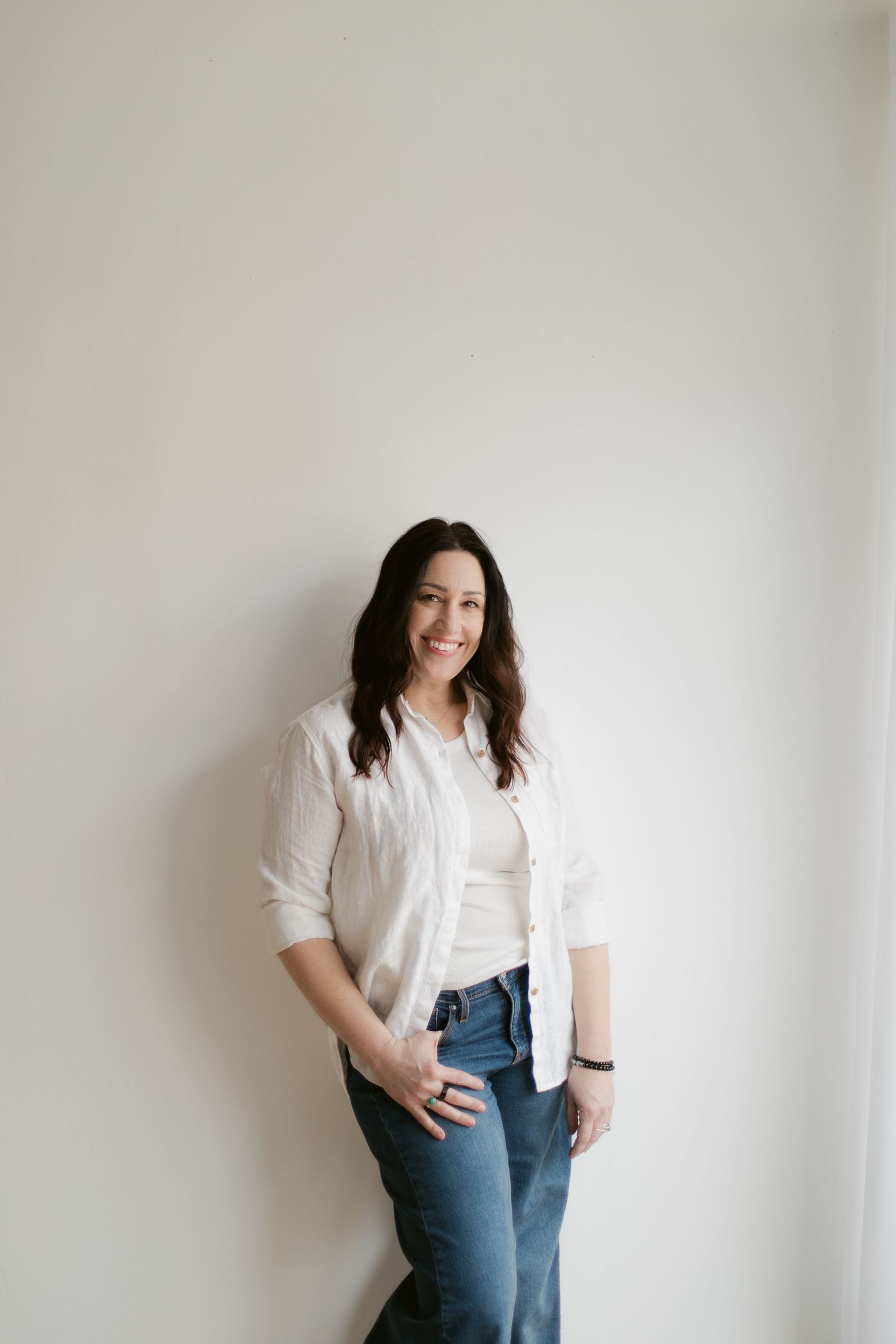 A smiling brunette woman in a white shirt and jeans leans against a light wall in a bright, minimalist setting.