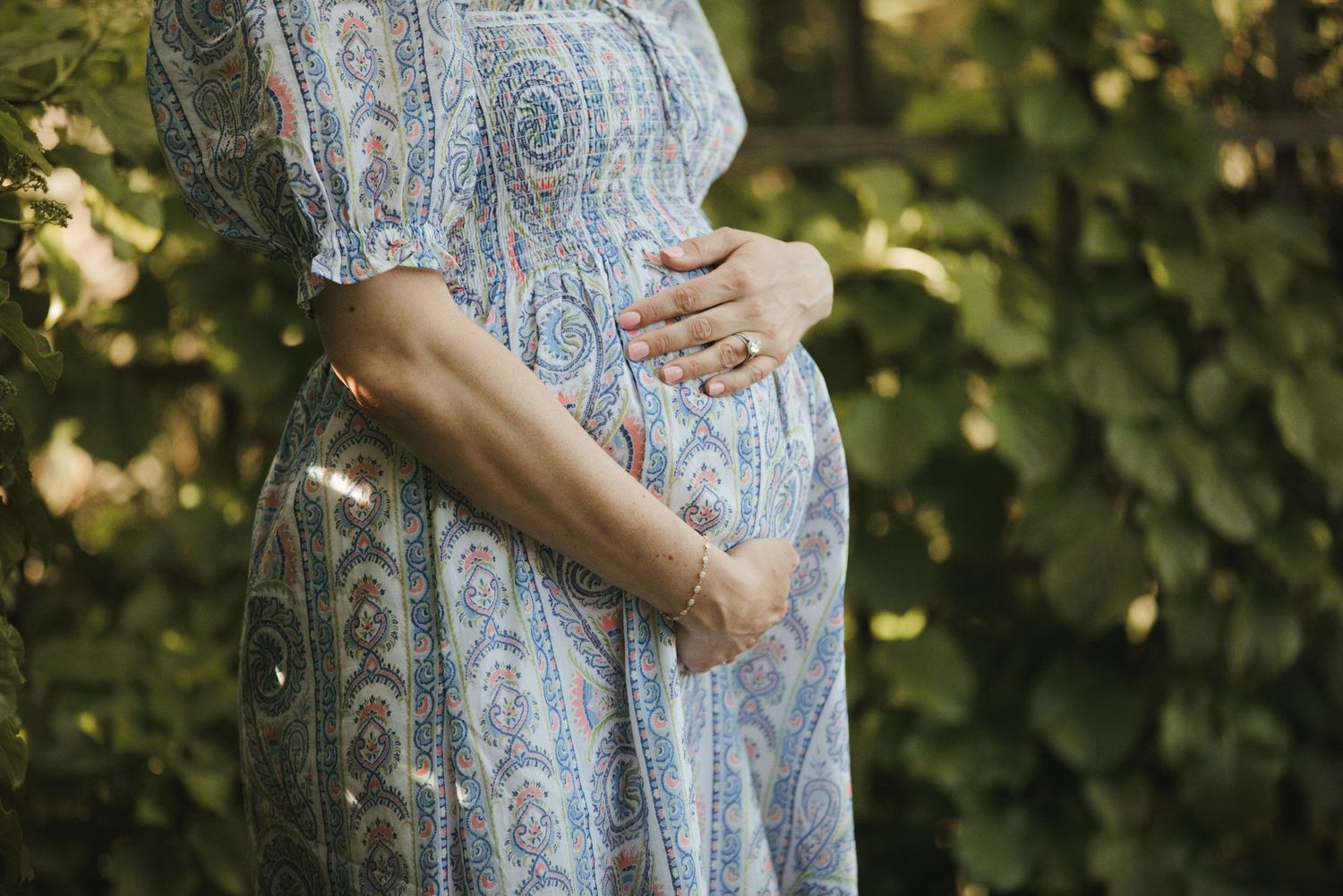 Close-up of pregnant woman in blue paisley dress cradling baby bump outdoors among green foliage.