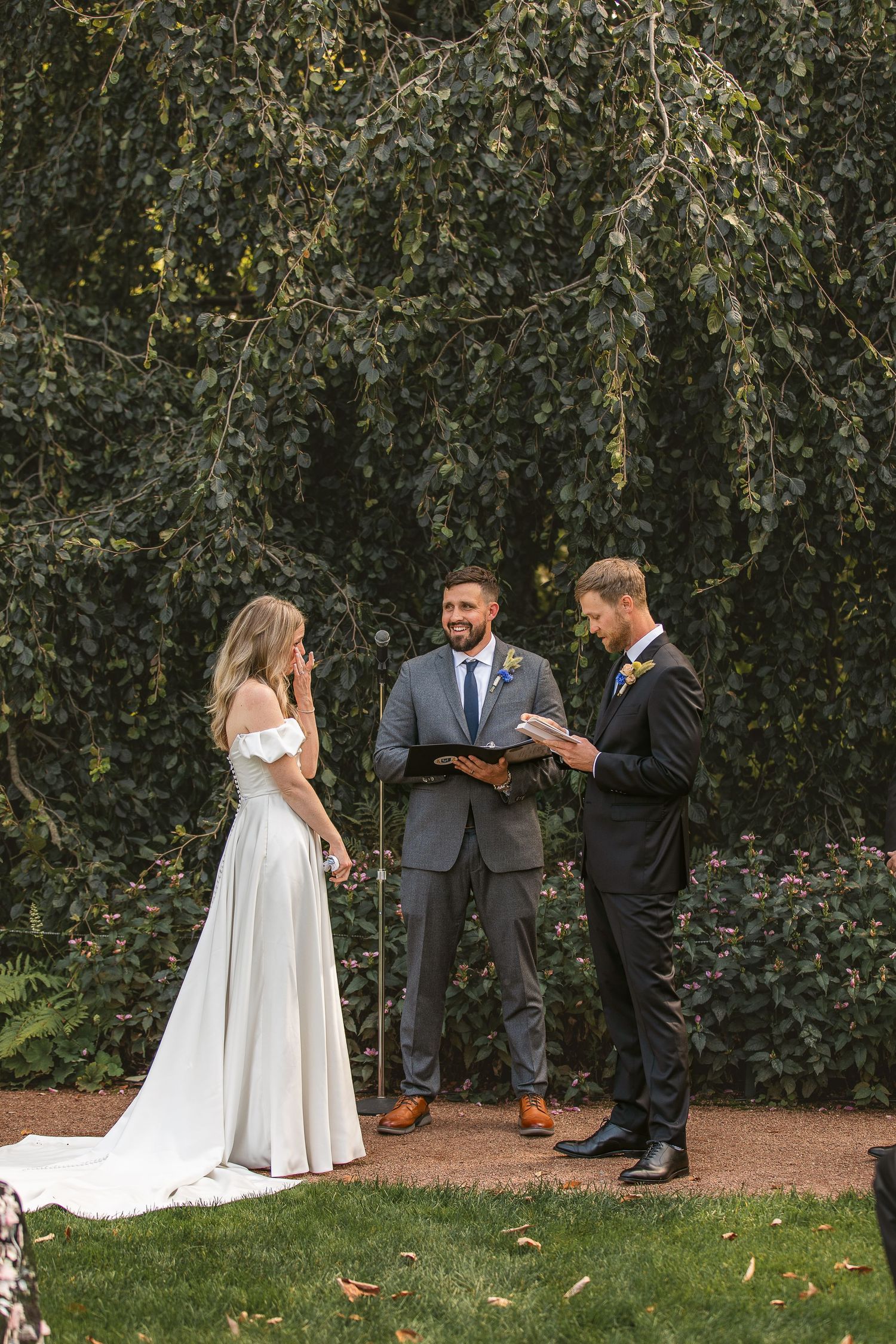 Wedding party members stand together on grass near hedge wall during outdoor autumn ceremony.