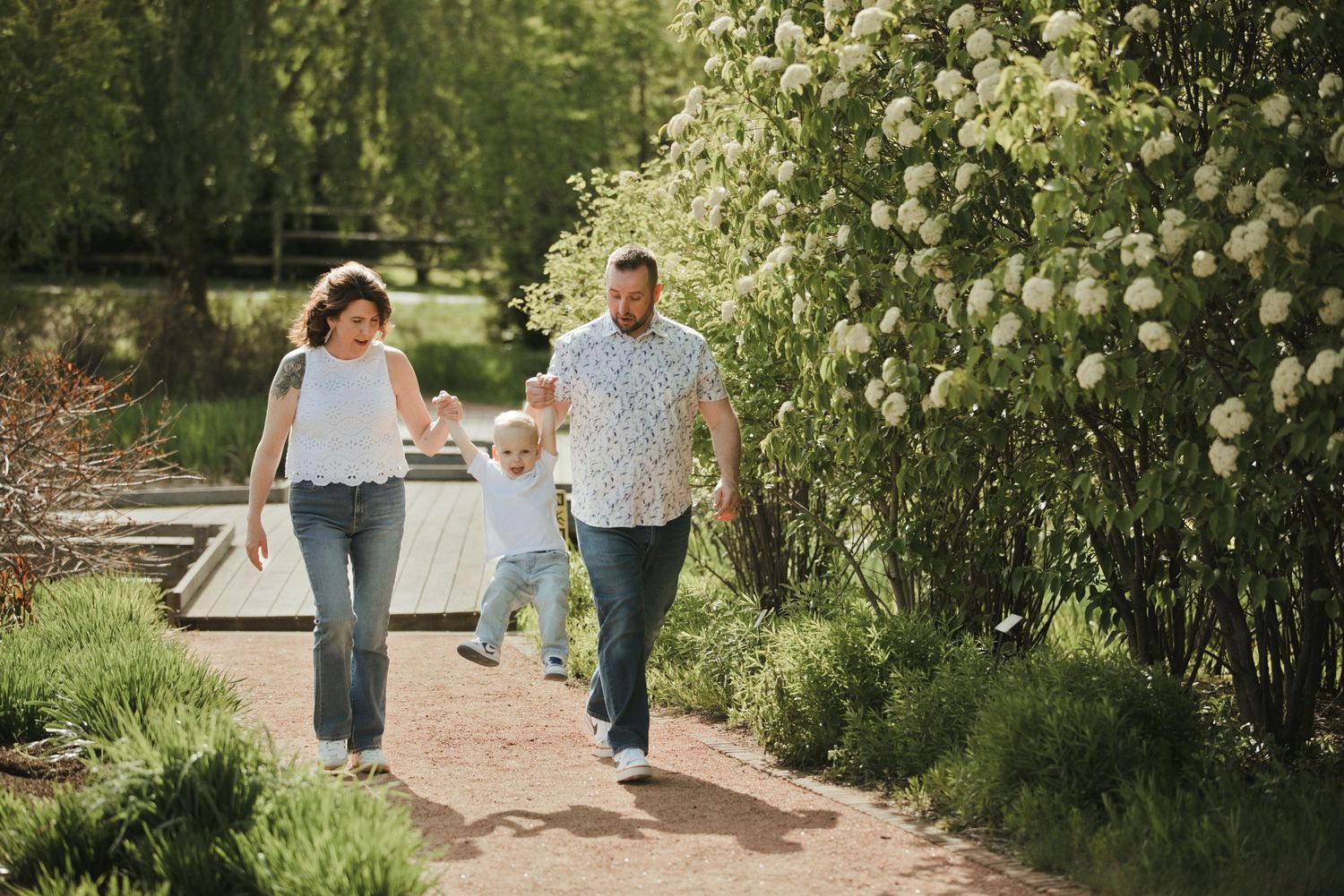 Family of three walking hand-in-hand on a garden path surrounded by lush greenery and white blooming flowers.
