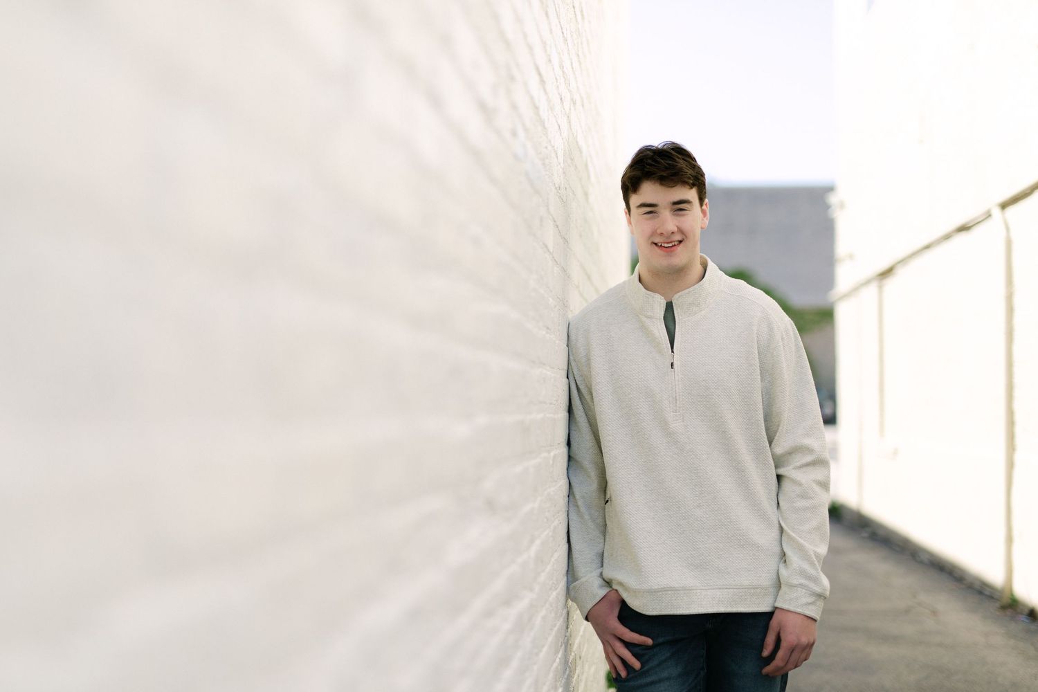 Young man in gray quarter-zip sweater smiling while leaning against a white brick wall outdoors.