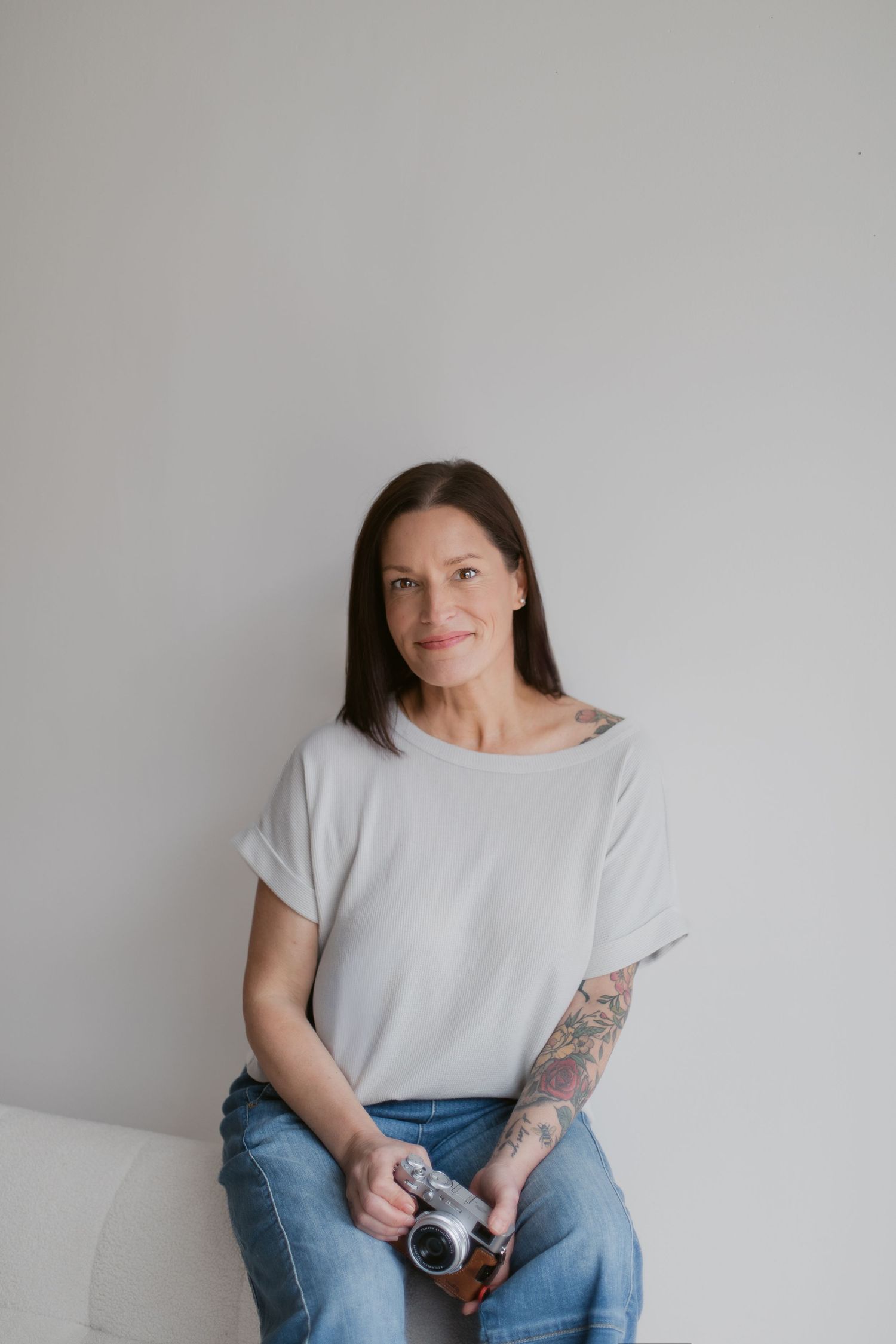 A person in a casual grey t-shirt and jeans sits on a white couch holding a vintage camera.