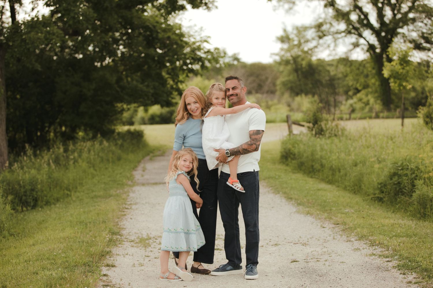 Family of four posing on a gravel path surrounded by lush green trees during a golden hour outdoor photo session.